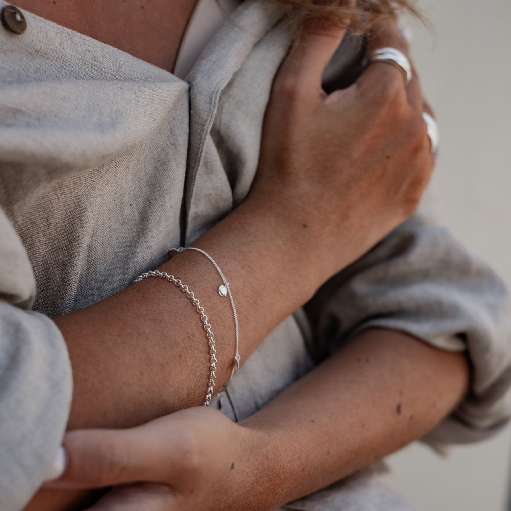 A woman with crossed arms, wearing layered silver bracelets with small circular charms and silver rings.