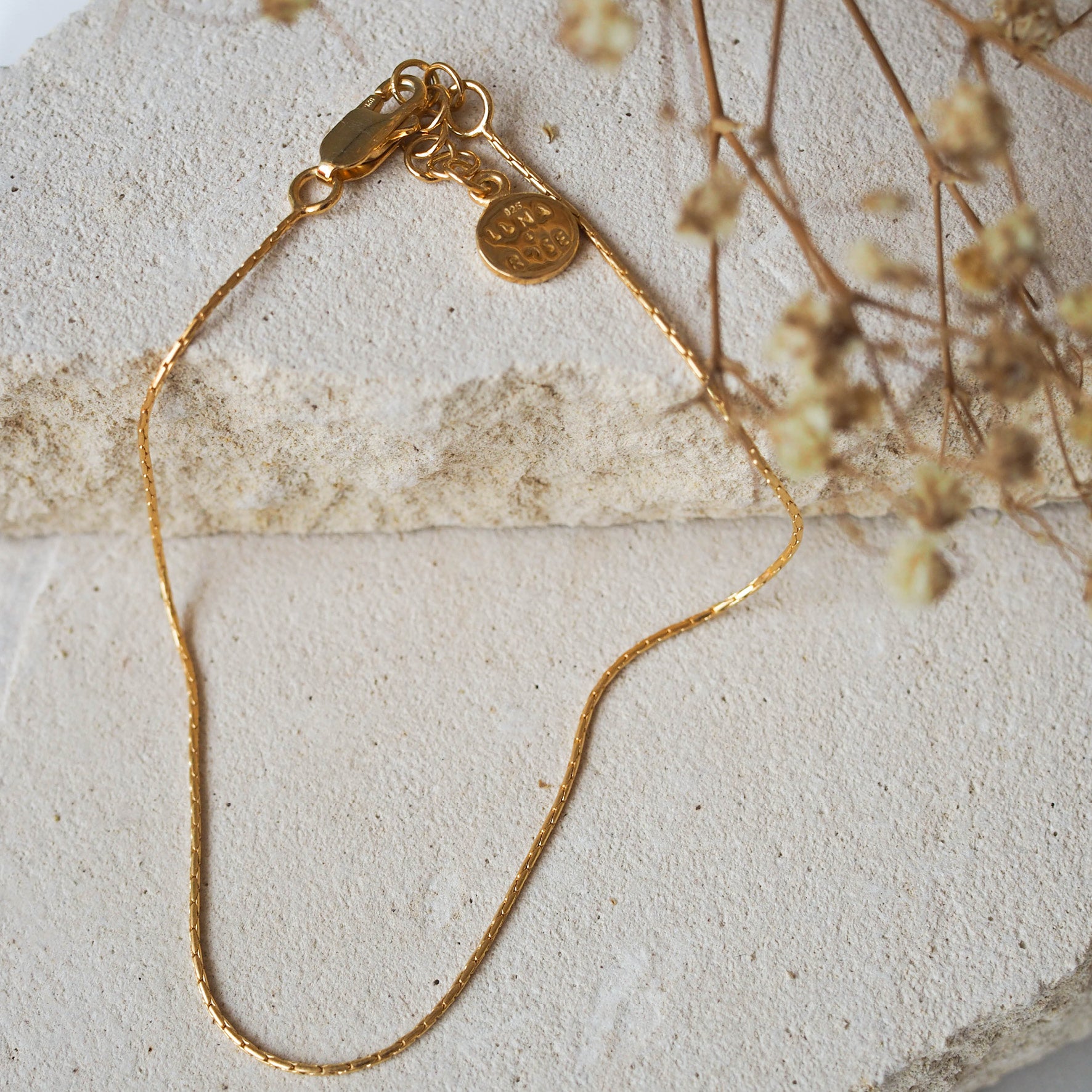 A delicate gold chain anklet, displayed on beige stone with dried flowers.