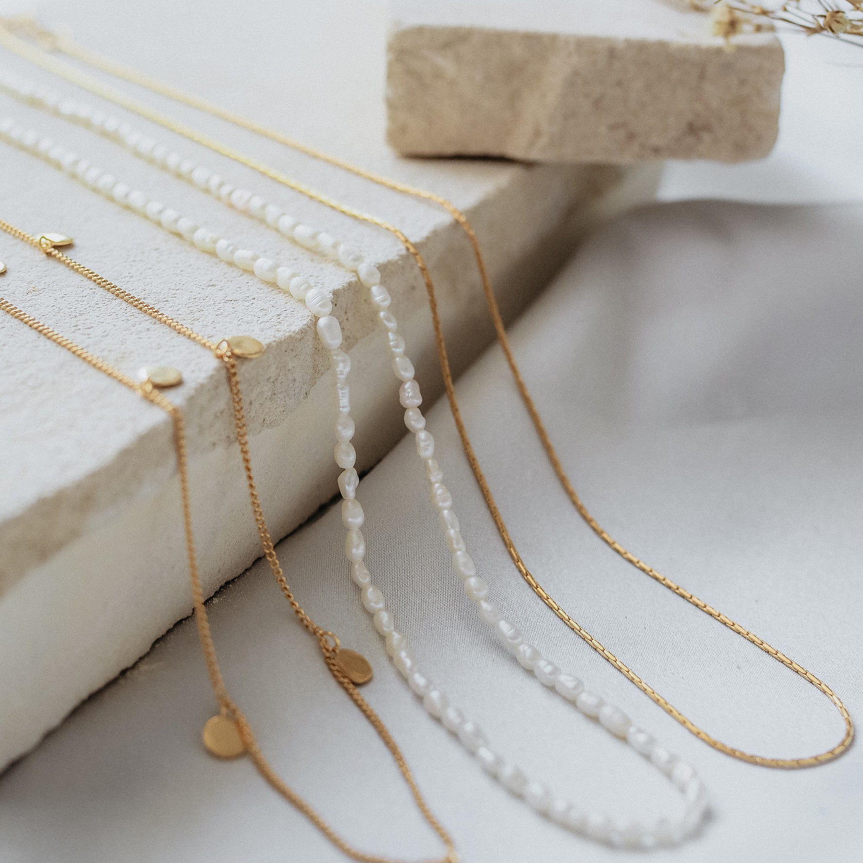 A display of delicate necklaces, including gold chains and pearl strands, arranged on beige stone with dried flowers.