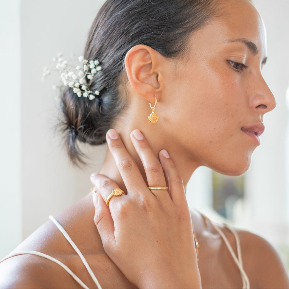 A close-up of a woman with gold sunburst earrings and gold rings, with flowers in her hair.