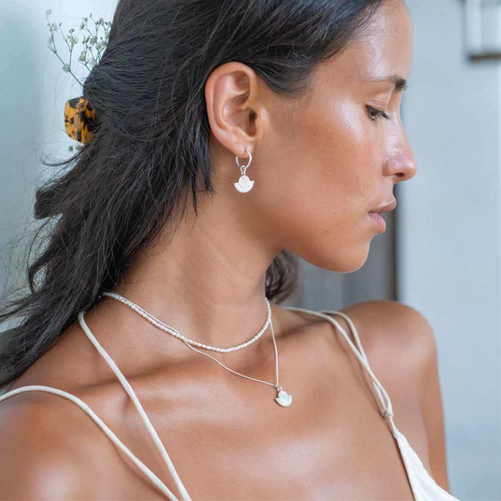 A close-up of a woman wearing a brown hair clip, silver sunburst earrings, a silver necklace with a sunburst pendant, and a pearl necklace.