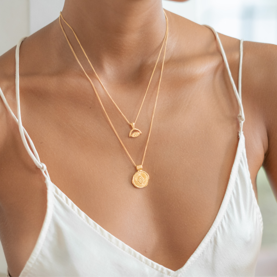 Close-up of layered gold necklaces with intricate pendants on a woman's neck, who is wearing a white strappy top.