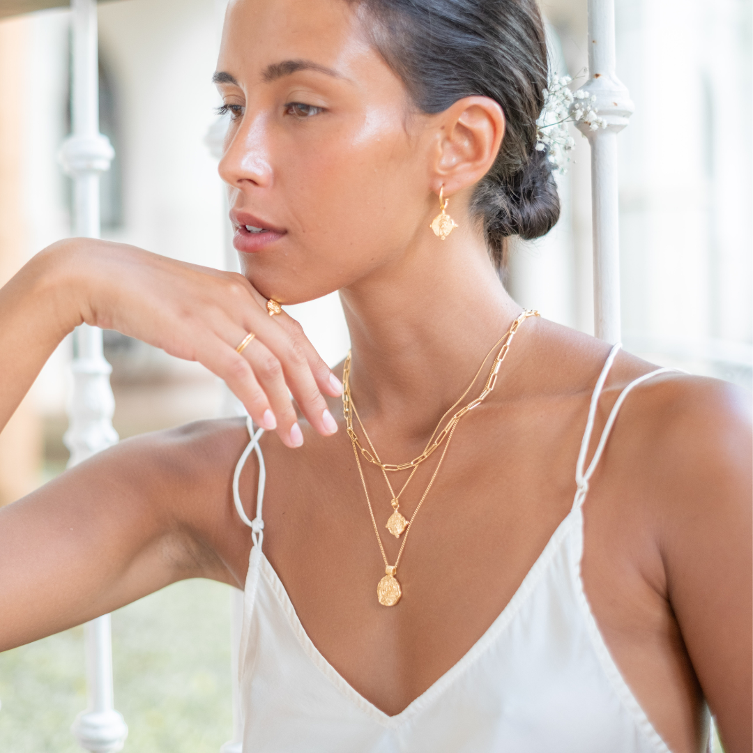 A woman wearing gold earrings, layered gold necklaces, and a white strappy top, with her hair styled in a bun adorned with small flowers.