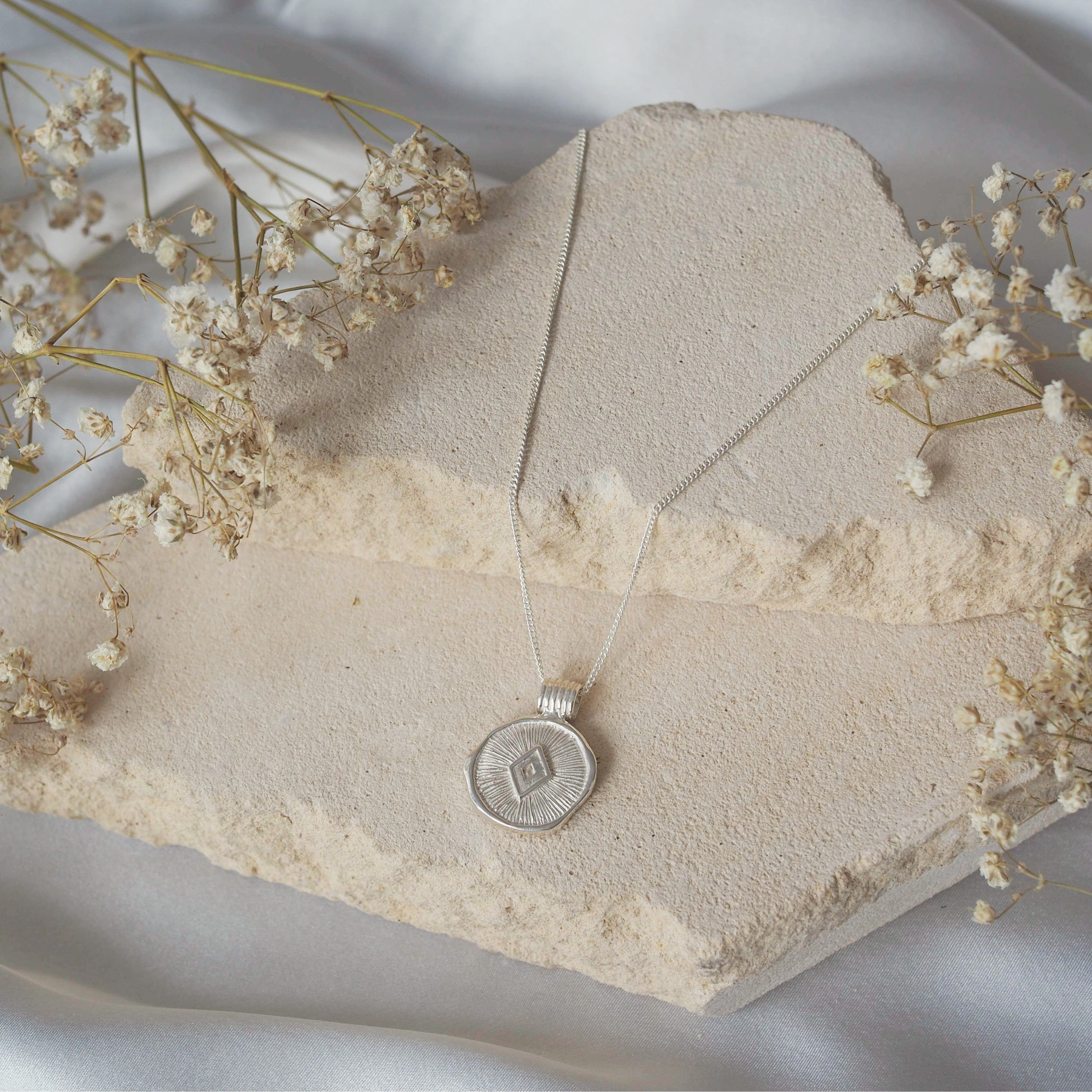 A silver necklace with a coin-shaped pendant featuring an etched diamond design, displayed on beige stone with dried flowers.