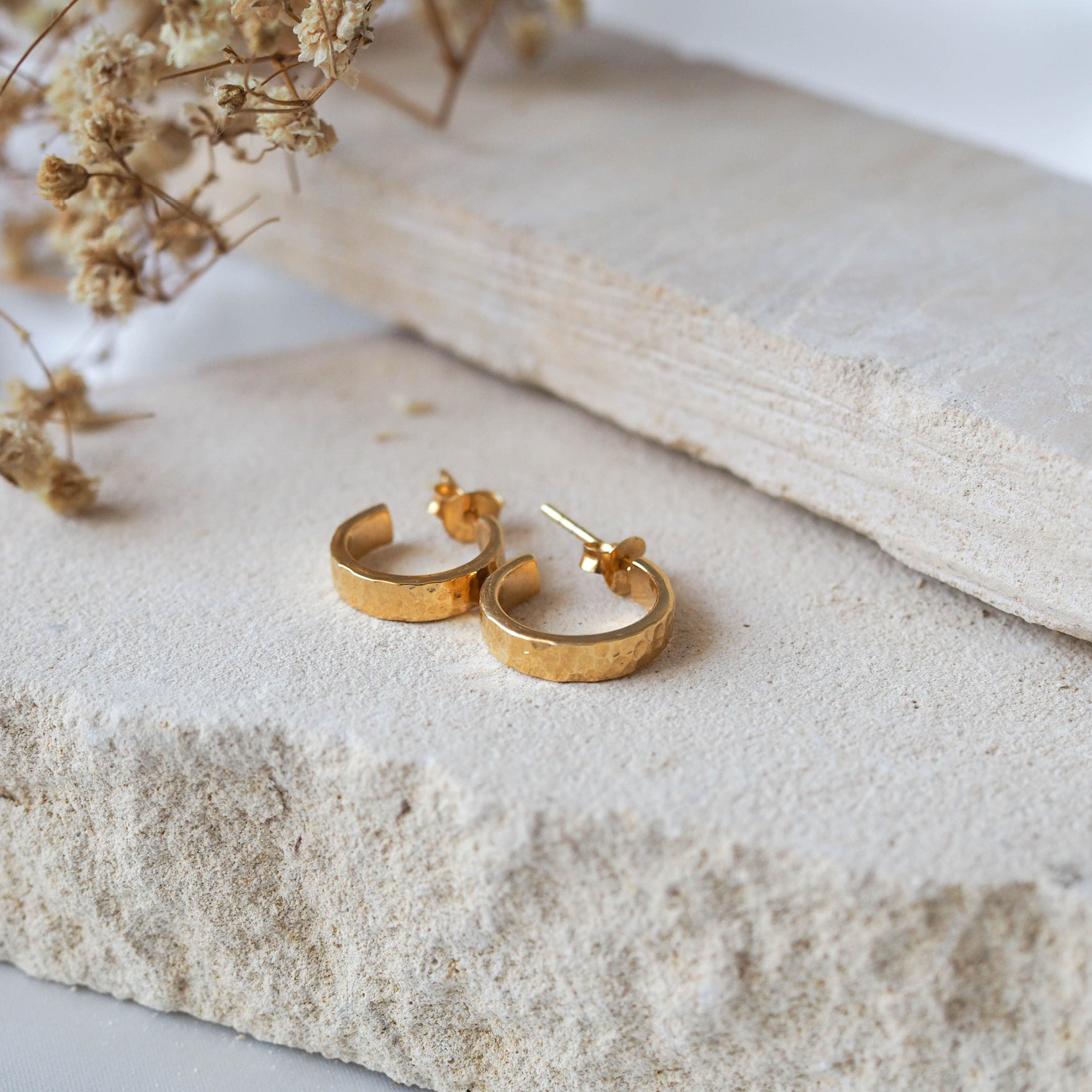 A pair of hammered gold hoop earrings displayed on textured stone beside dried flowers.