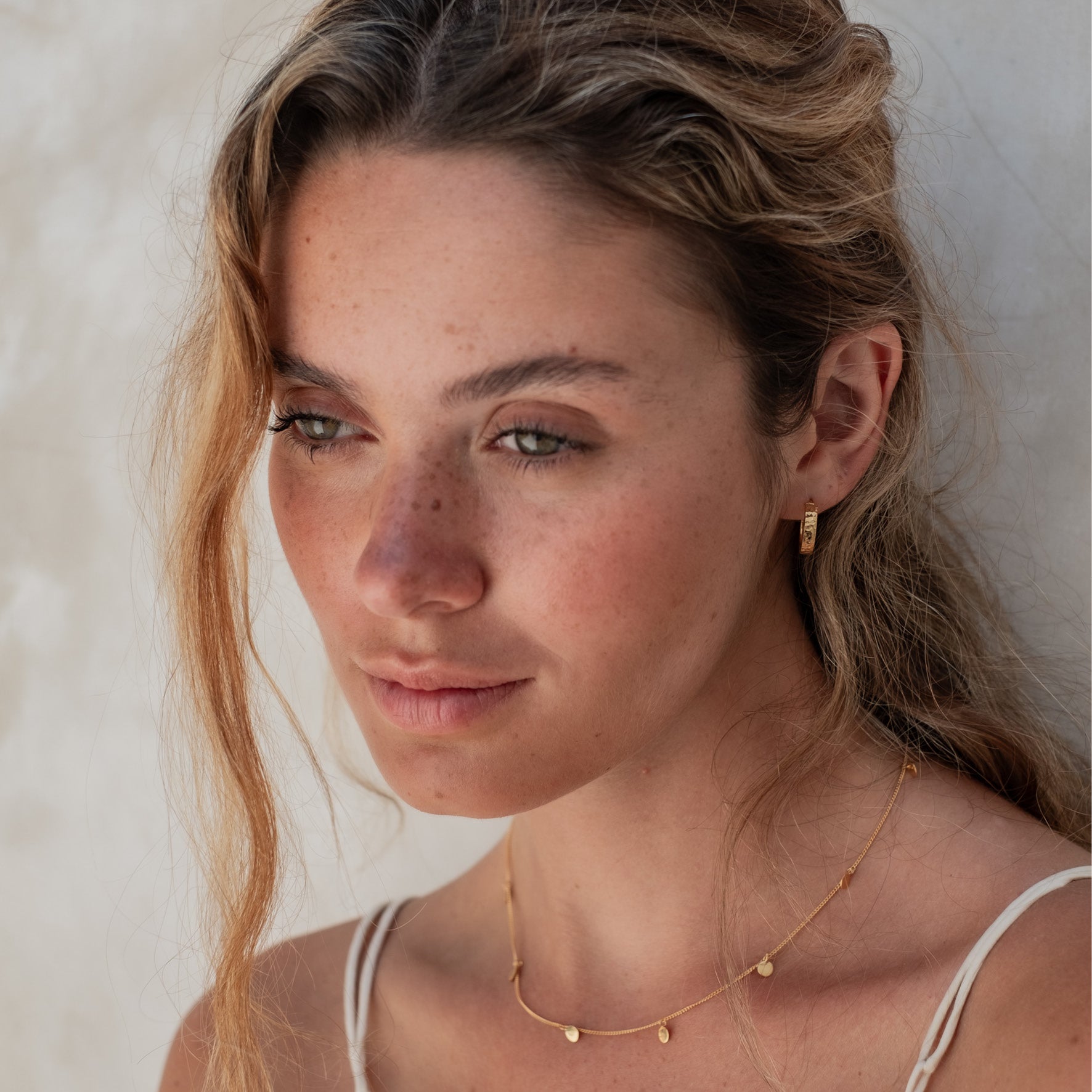 A woman wearing a hammered gold hoop earring and a delicate gold necklace with charms, looking away.