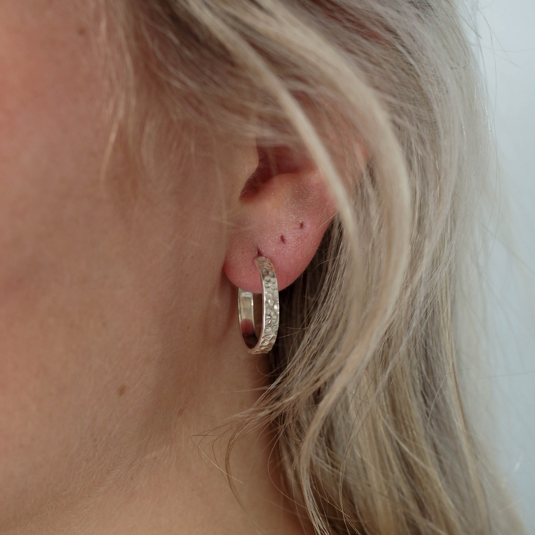 Close-up of a single hammered silver hoop earring worn on an ear with blonde hair.