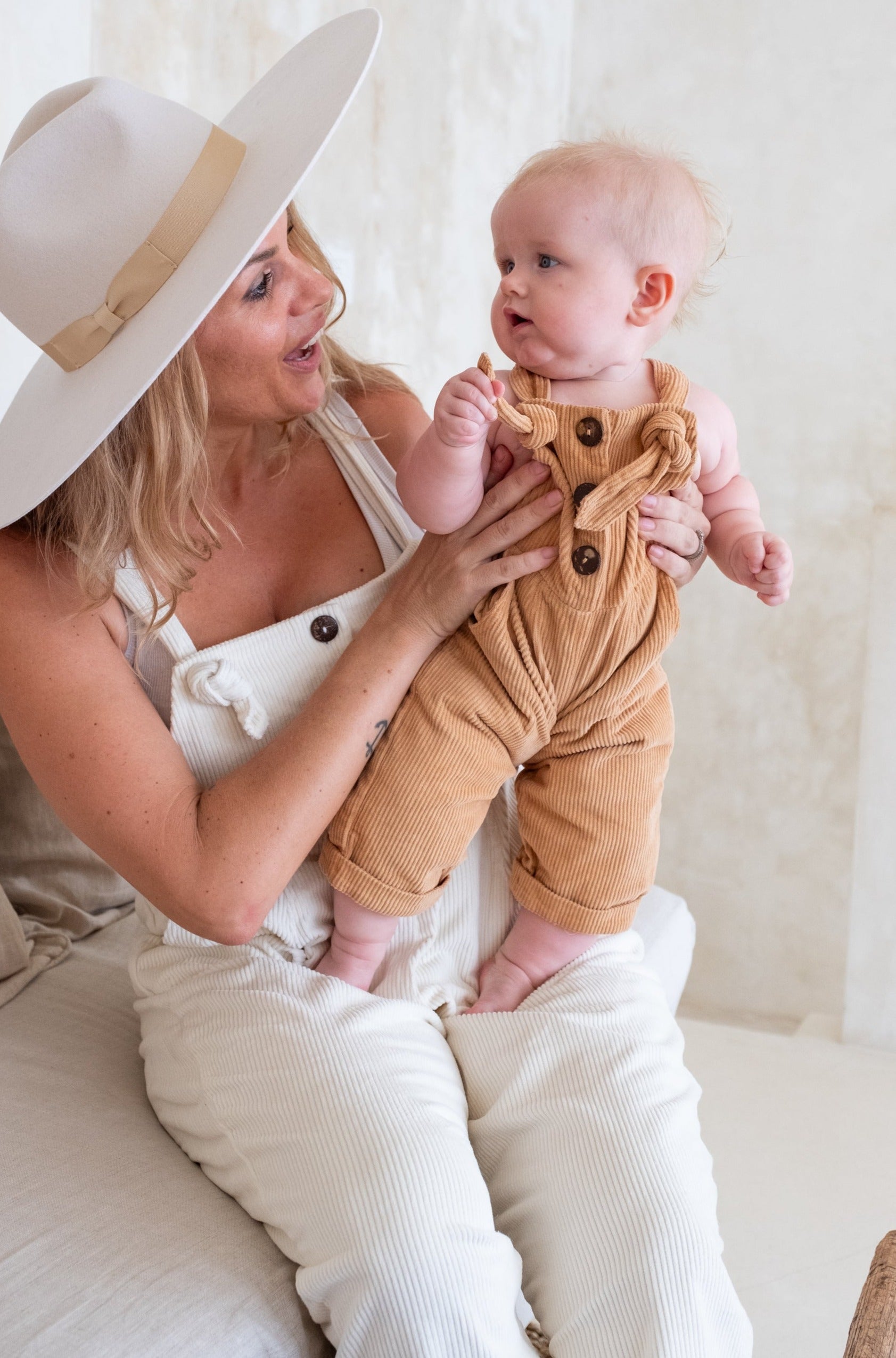 A woman in white overalls and a wide-brimmed hat holds up a baby dressed in Cinnamon overalls.