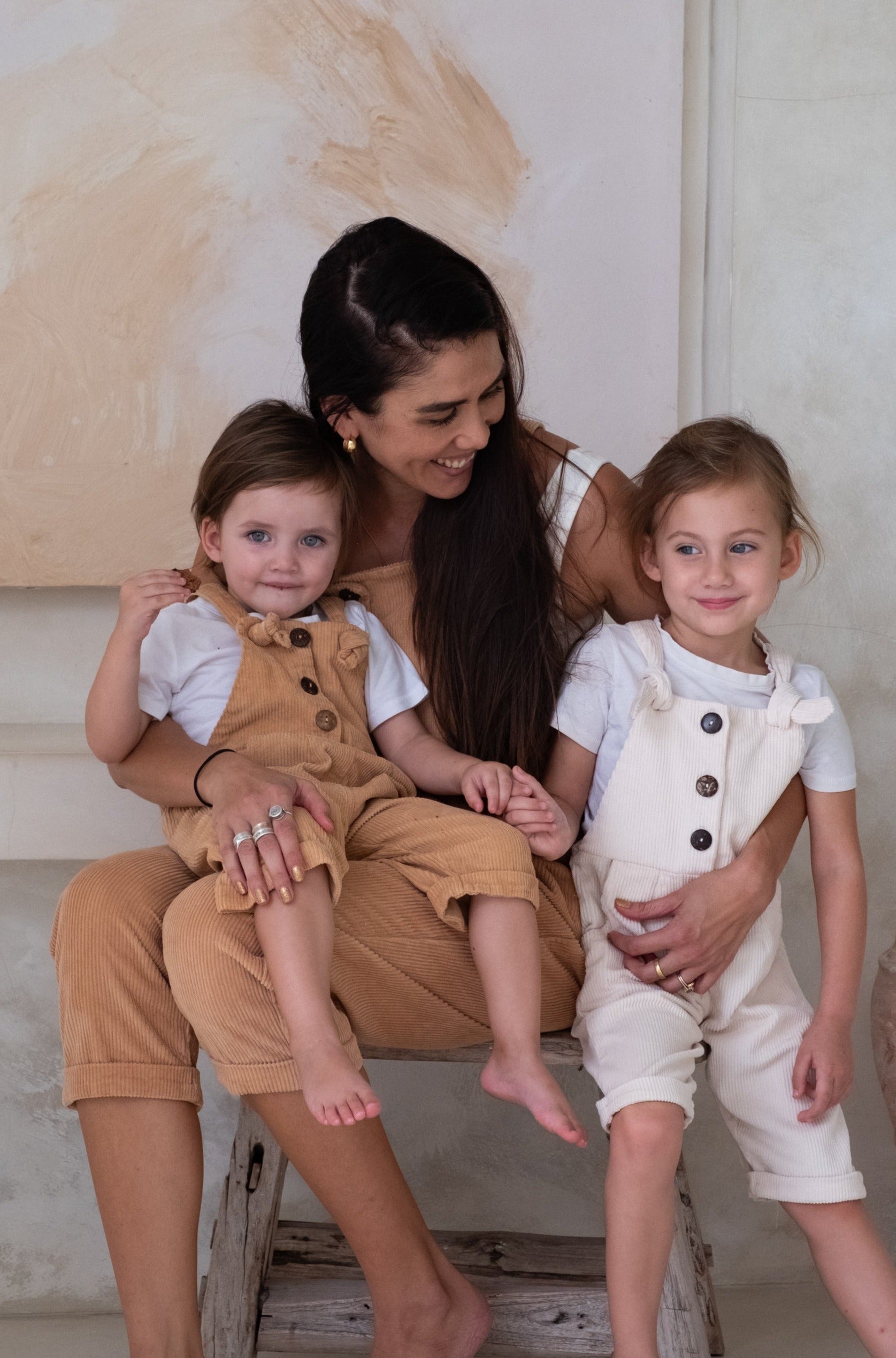 A woman in tan overalls sits on a bench with two young girls dressed in brown and white overalls.