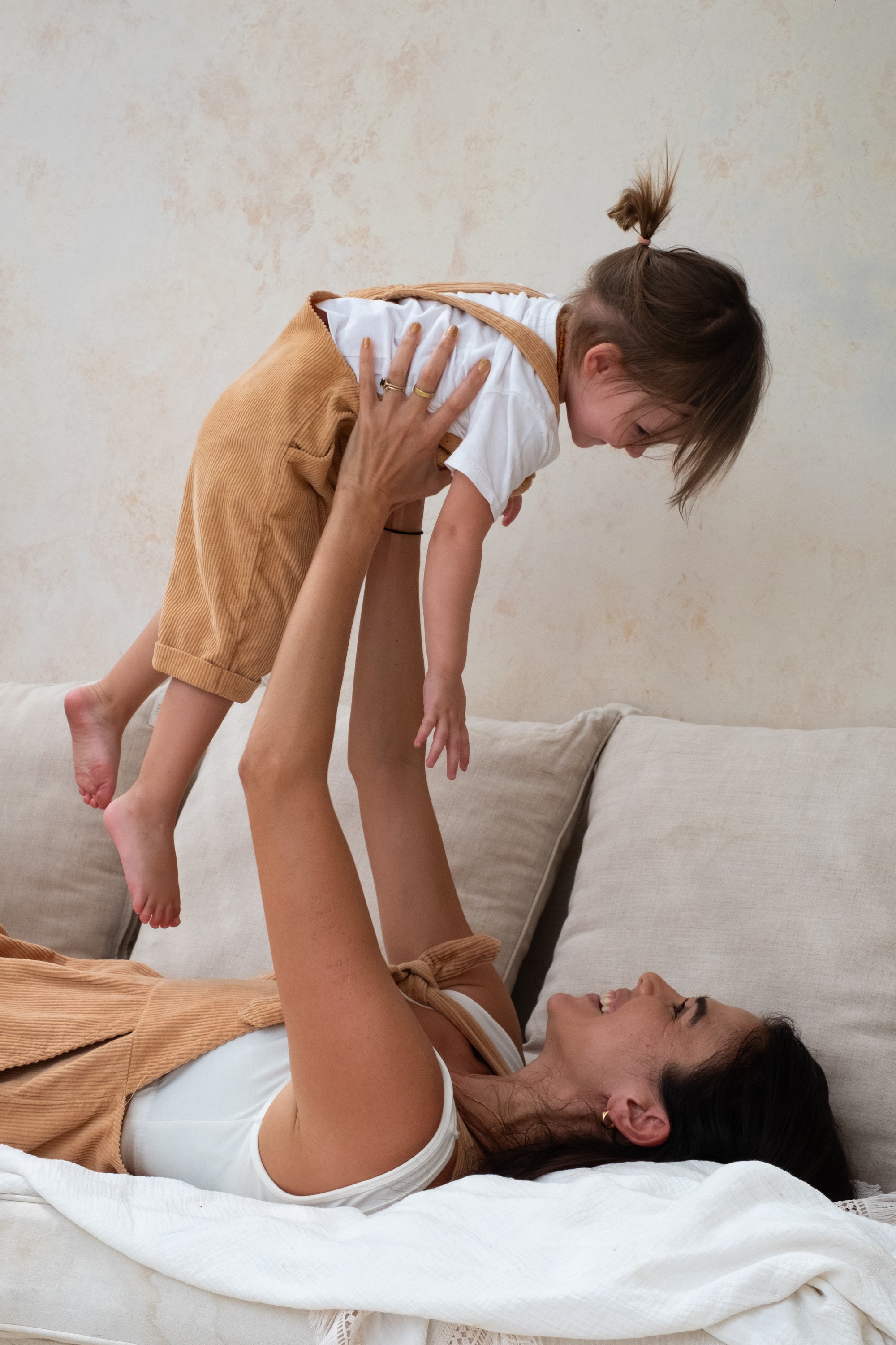 A woman lying on a beige couch playfully lifts a toddler dressed in tan overalls and a white shirt.