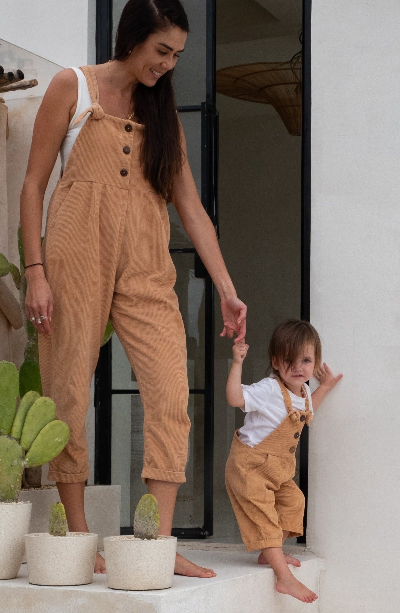 A woman in tan overalls holds hands with a toddler wearing tan overalls and a white shirt.