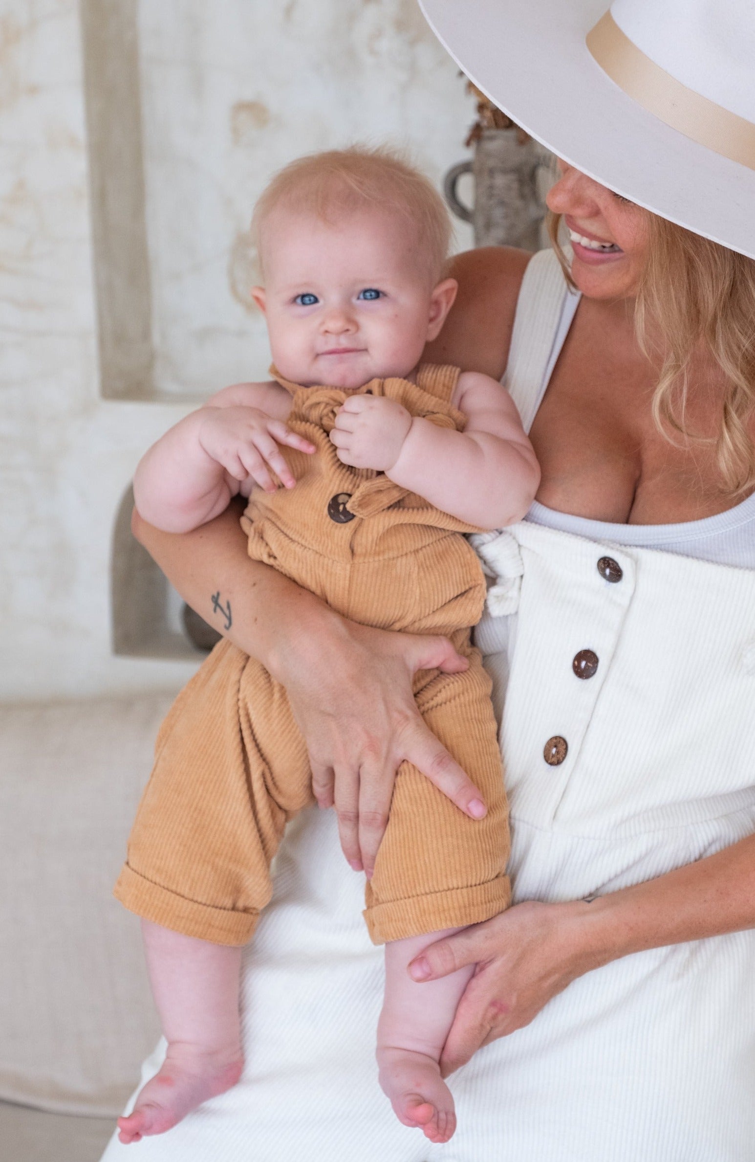 A smiling woman in white overalls with a wide-brimmed hat holds a baby in tan overalls.