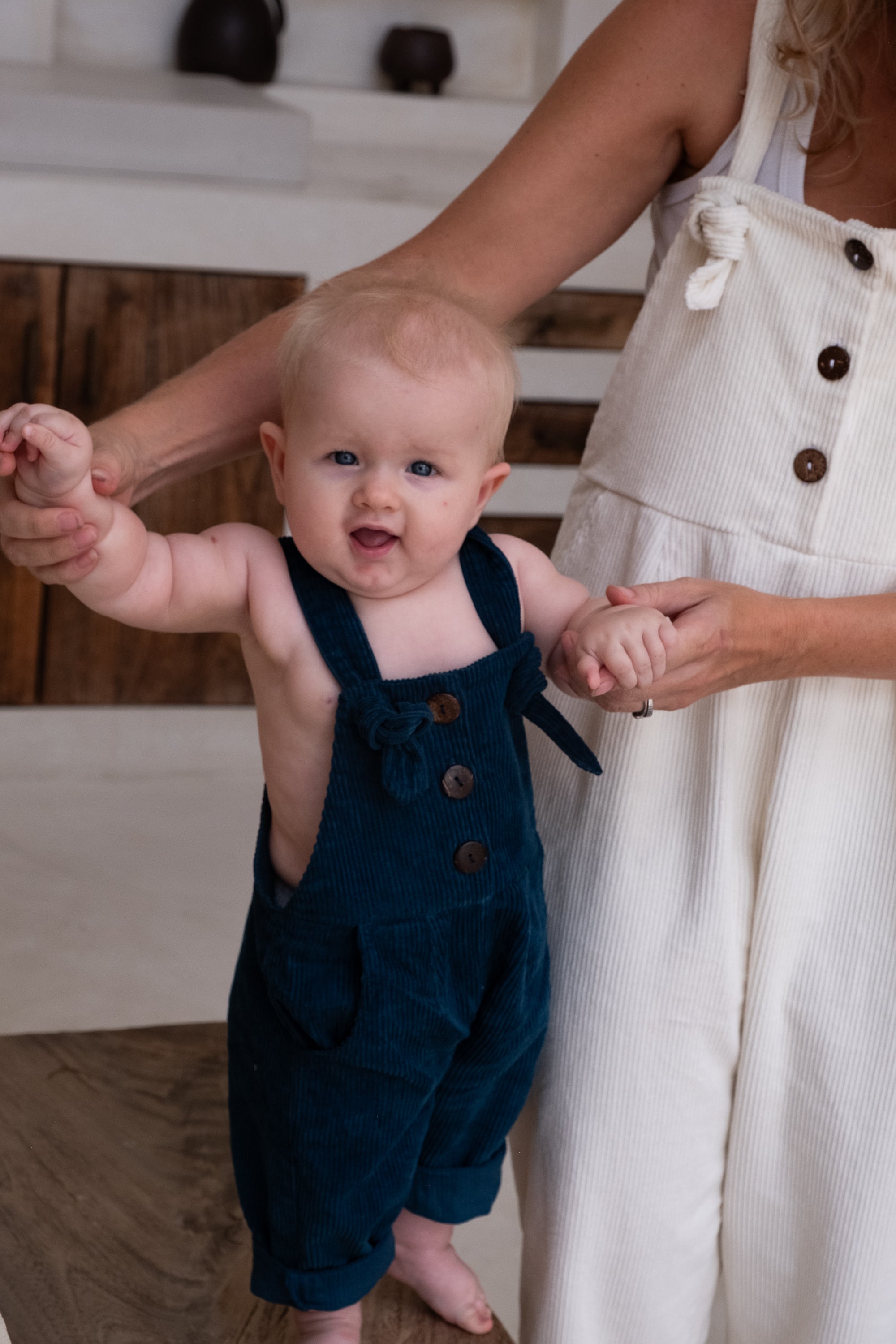 A close up of a baby in blue overalls stand on a wooden bench holding hands with a woman in white overalls