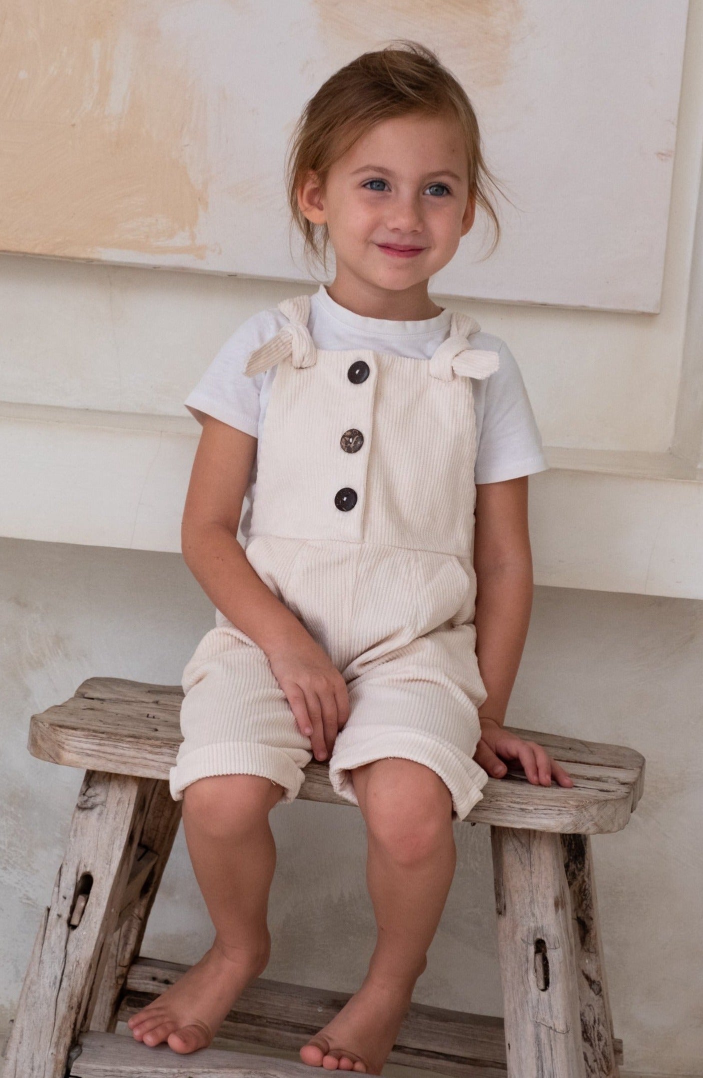 A child in a white overall and white t-shirt sitting on a wooden bench