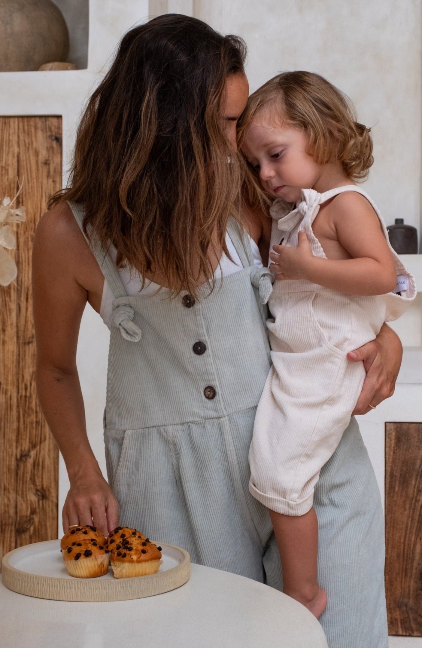 A child with her mother both wearing an overall staring at some cupcakes