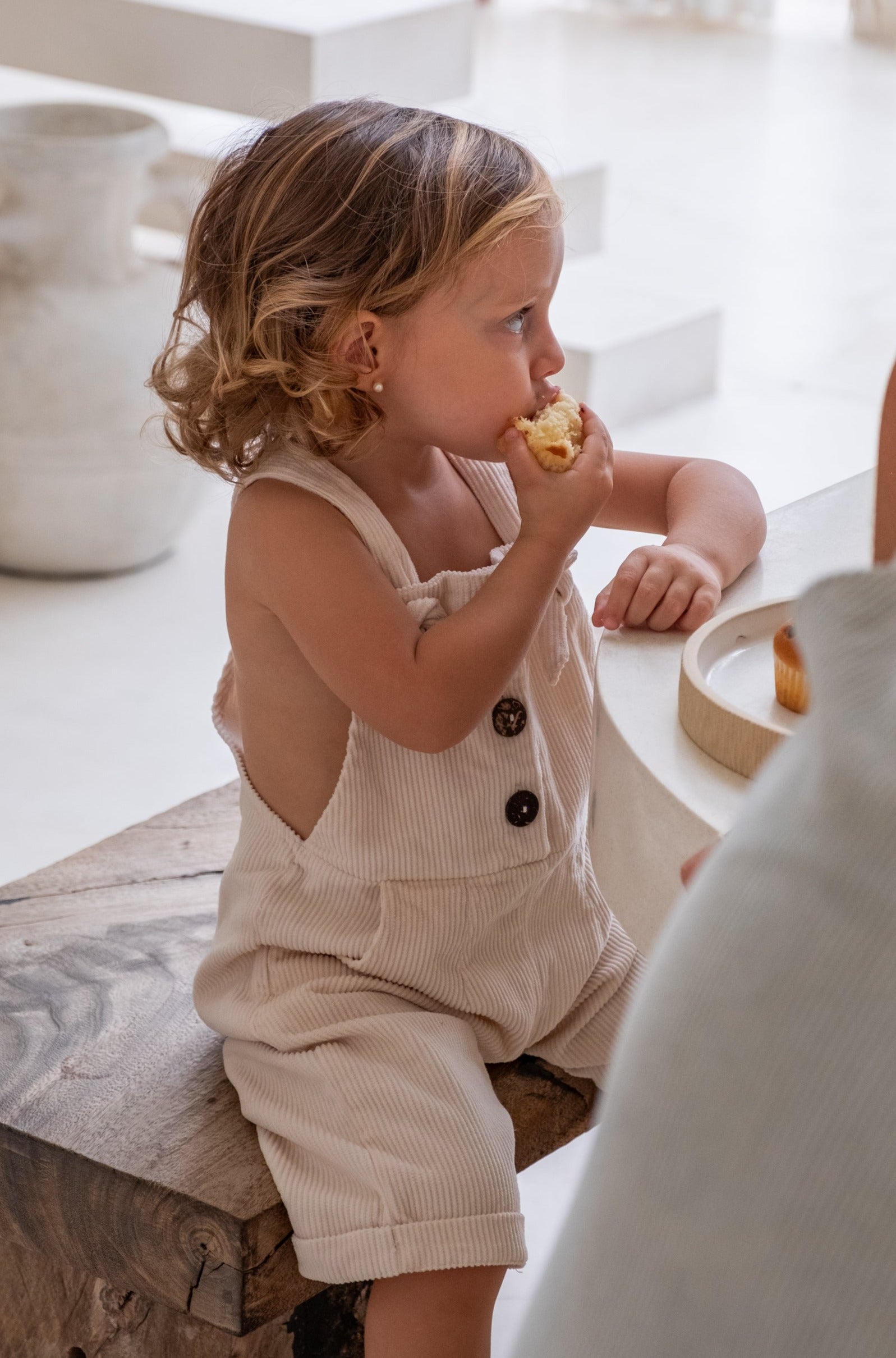 A toddler with blond her in a beige overall siting on a wooden bench eating a cupcake