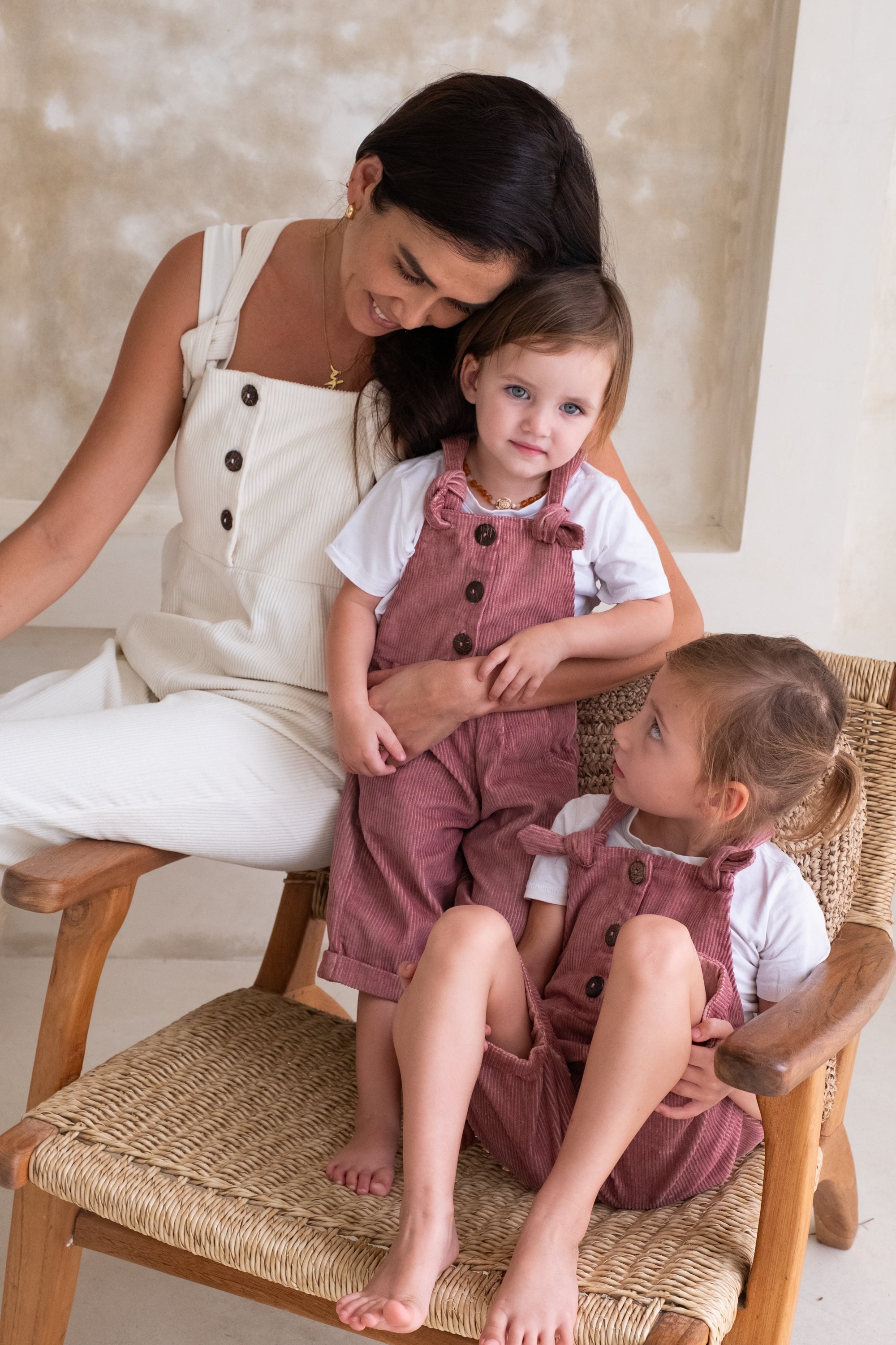A woman in a white overall together with two toddlers in a pink overall sitting on a chair