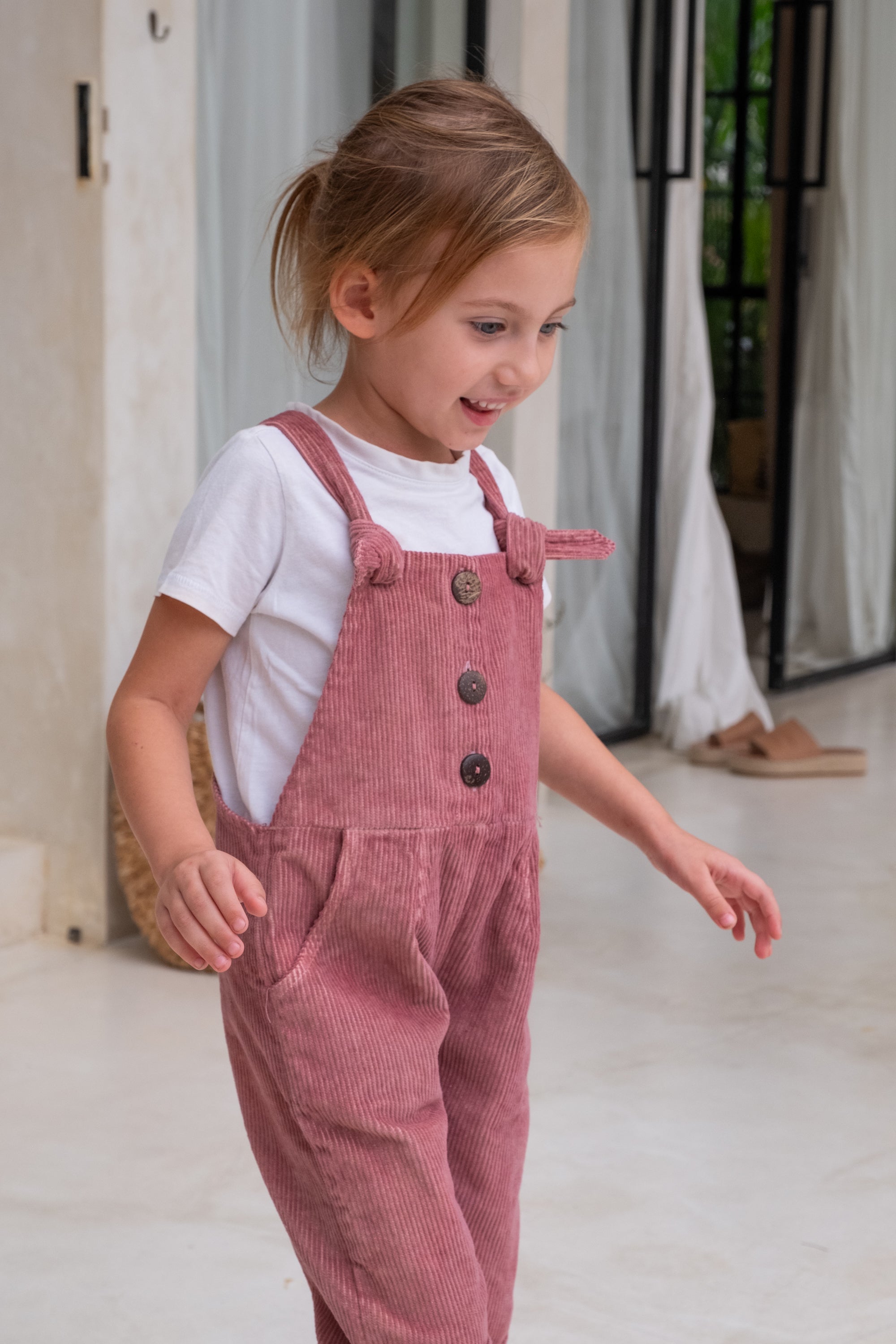 A child with brown hair and blue eyes smiling in her pink overall