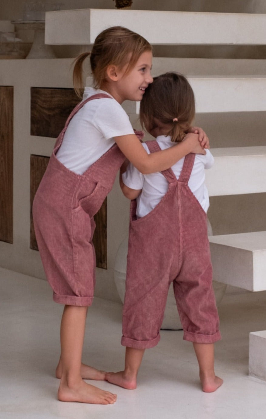 Two childs hugging each other while wearing a pink overall
