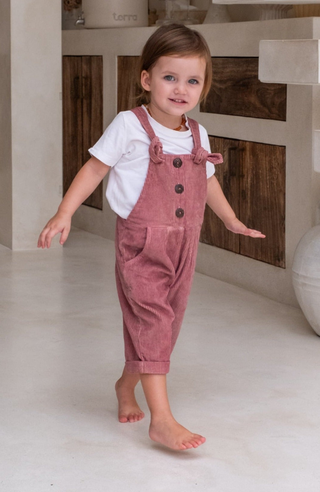 A toddler with blue eyes and brown hair walking in a pink overall