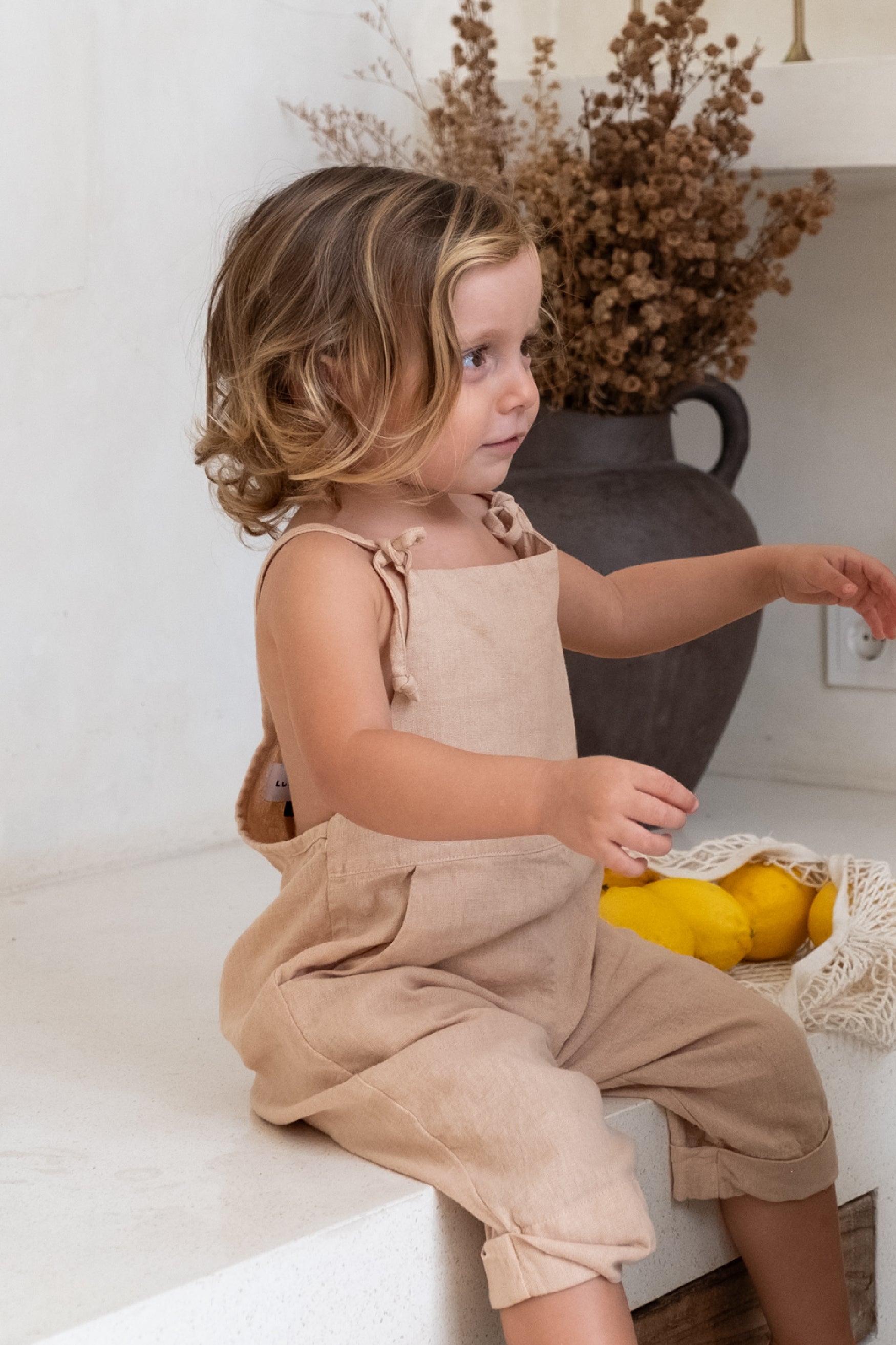 A toddler with blond hair sitting on top of the counter wearing a brown overal