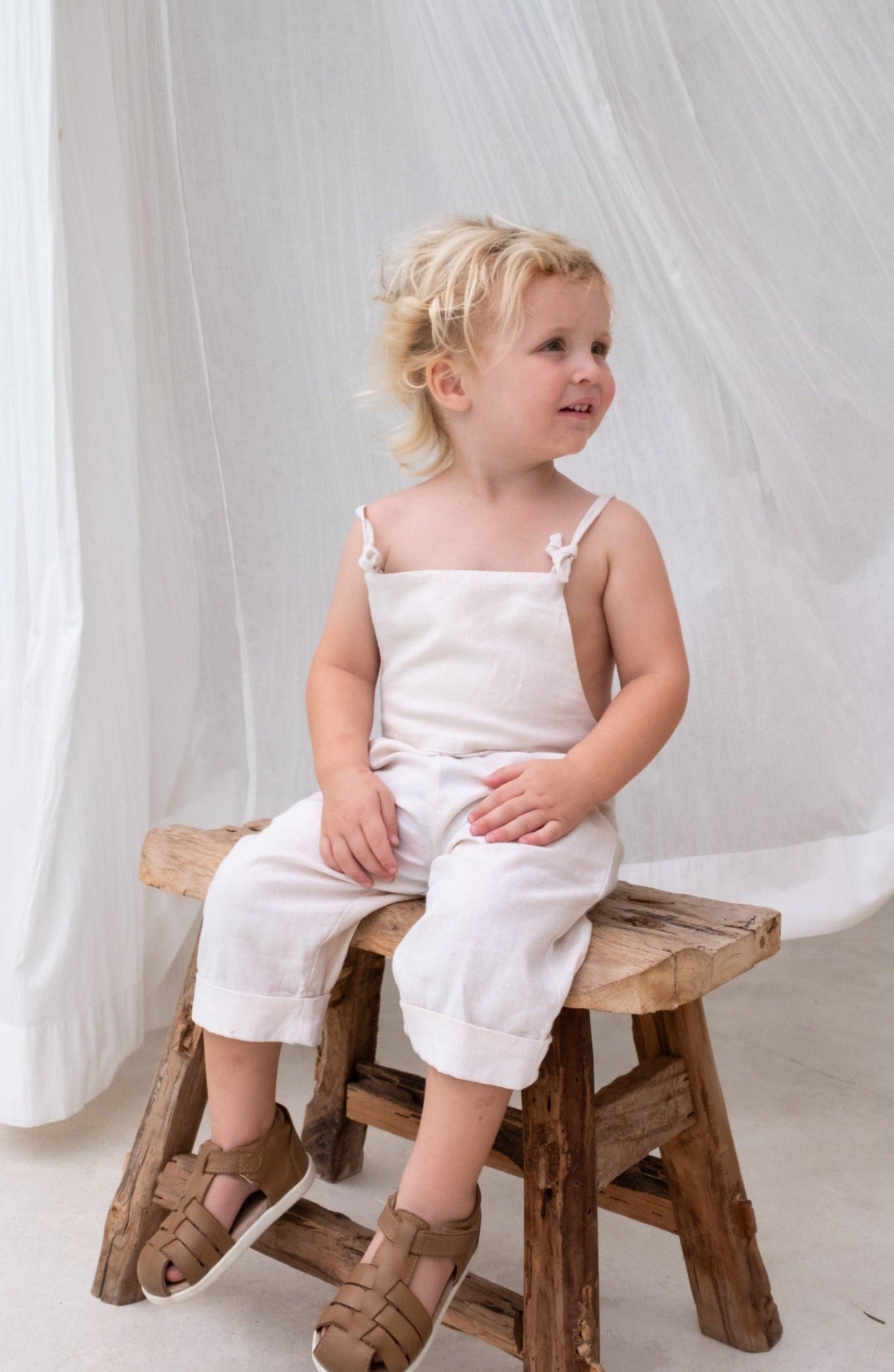 A child with blond hair sitting on a wooden stool wearing a white overall