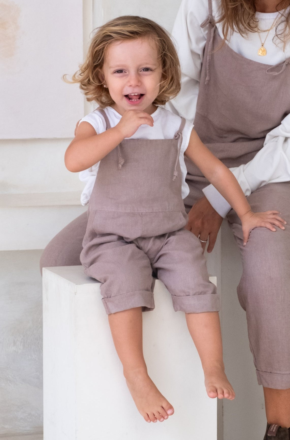 A toddler with brown hair sitting on a white object wearing a purple overall