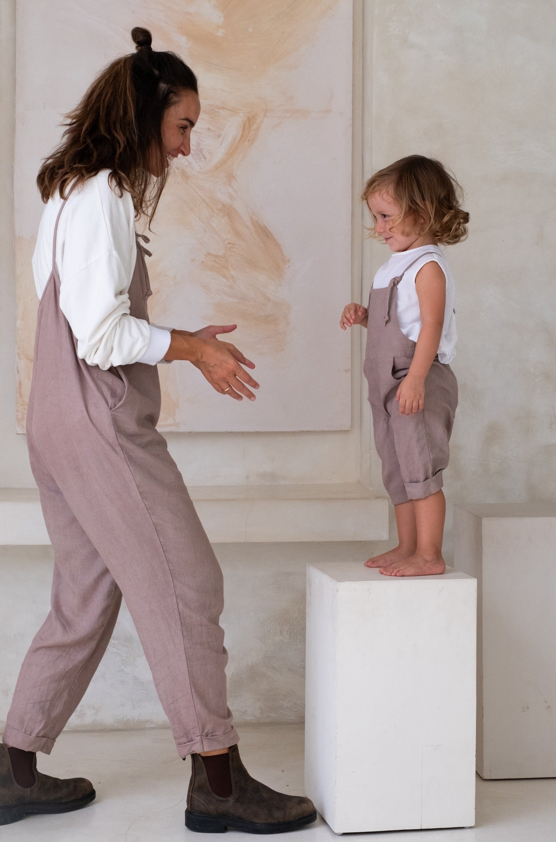 A woman in taupe overalls stands facing a toddler in matching overalls, who is standing on a white pedestal.