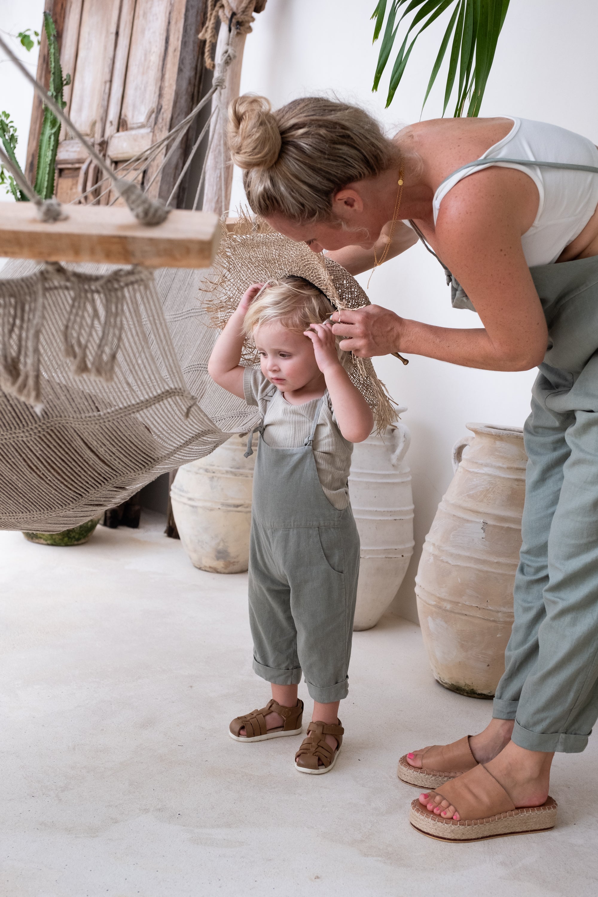 A toddler together with a woman both wearing an overall in the colour sage trying on a hat.