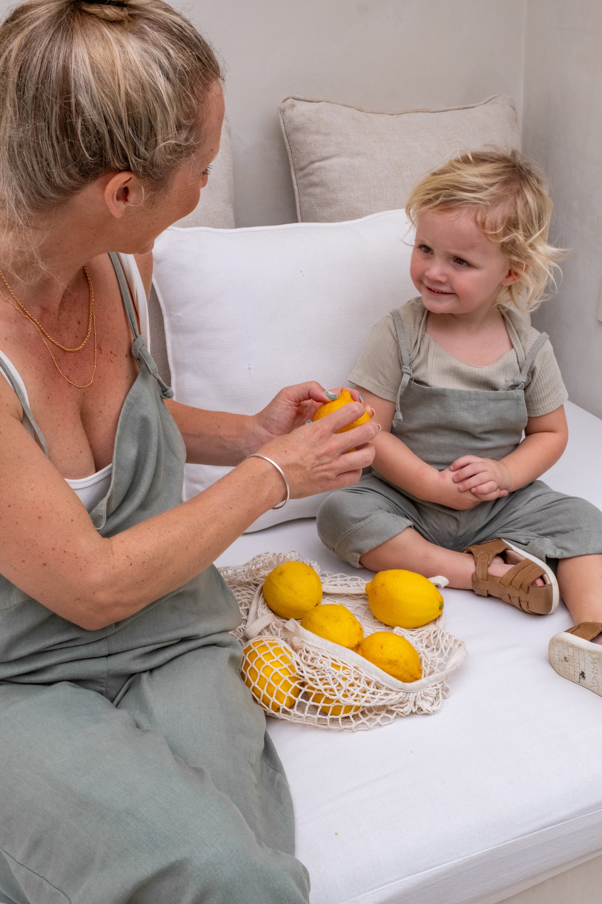 A toddler and a woman sitting on a couch both wearing an ovearll while looking into some lemons