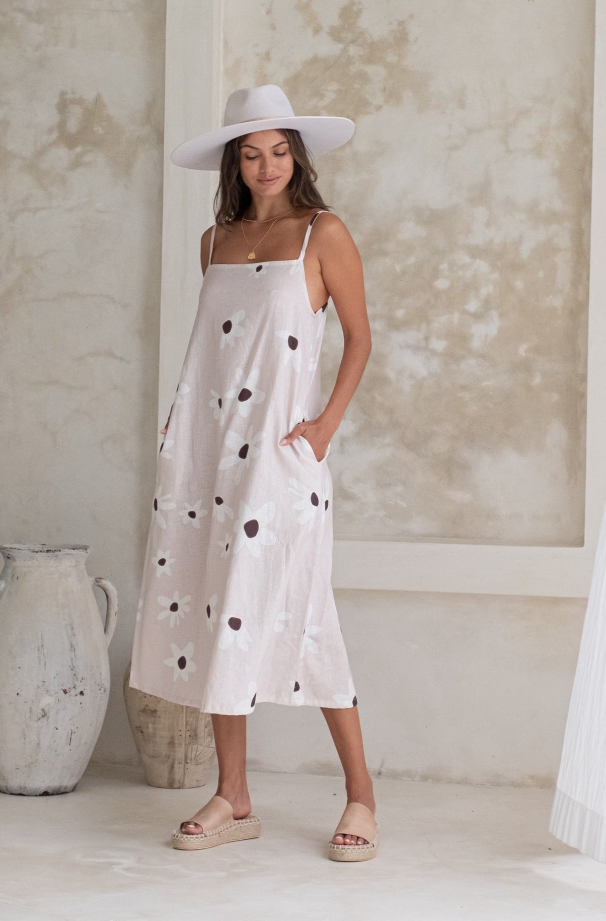 A woman in a floral sundress and wide-brimmed hat, looking down with her hands in her pockets, placed in a minimalistic indoor setting.