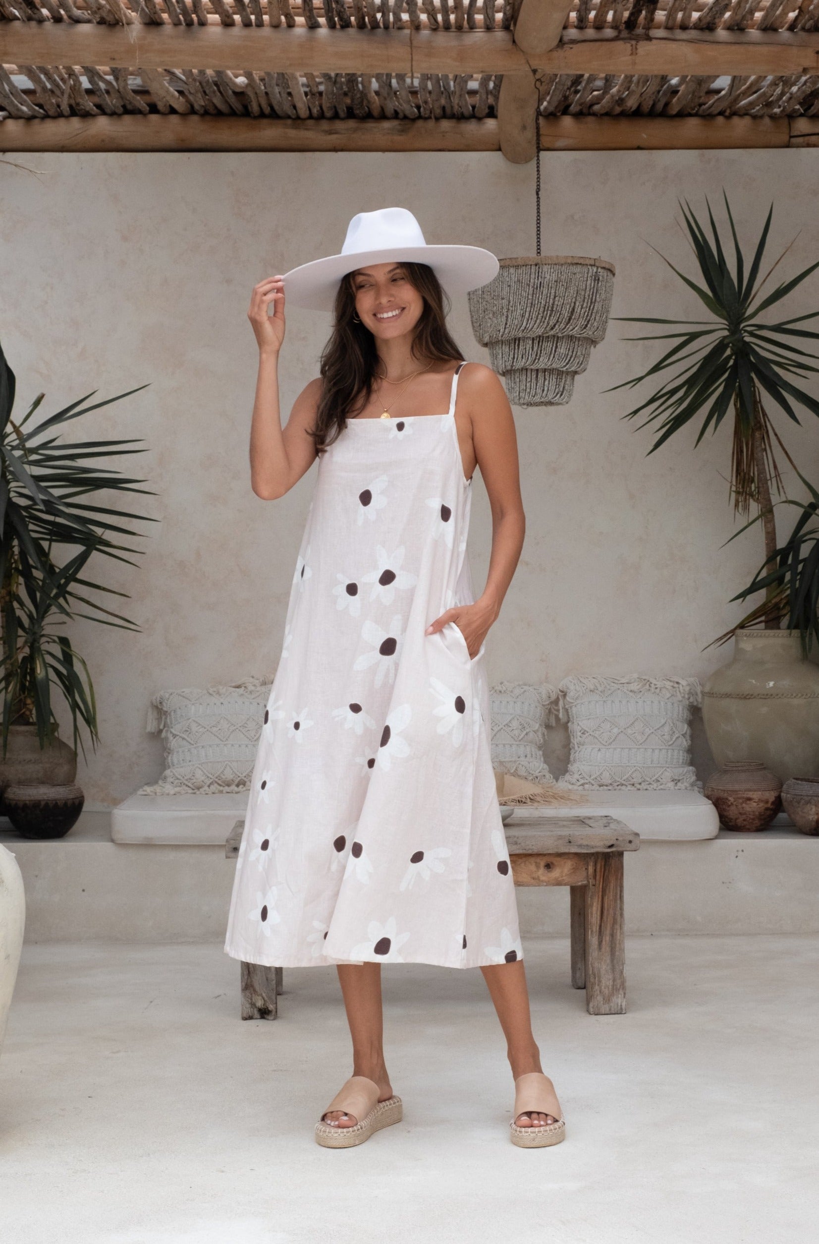 A woman wearing a white sundress adorned with floral patterns and a wide-brimmed hat, standing in a minimalistic outdoor setting.