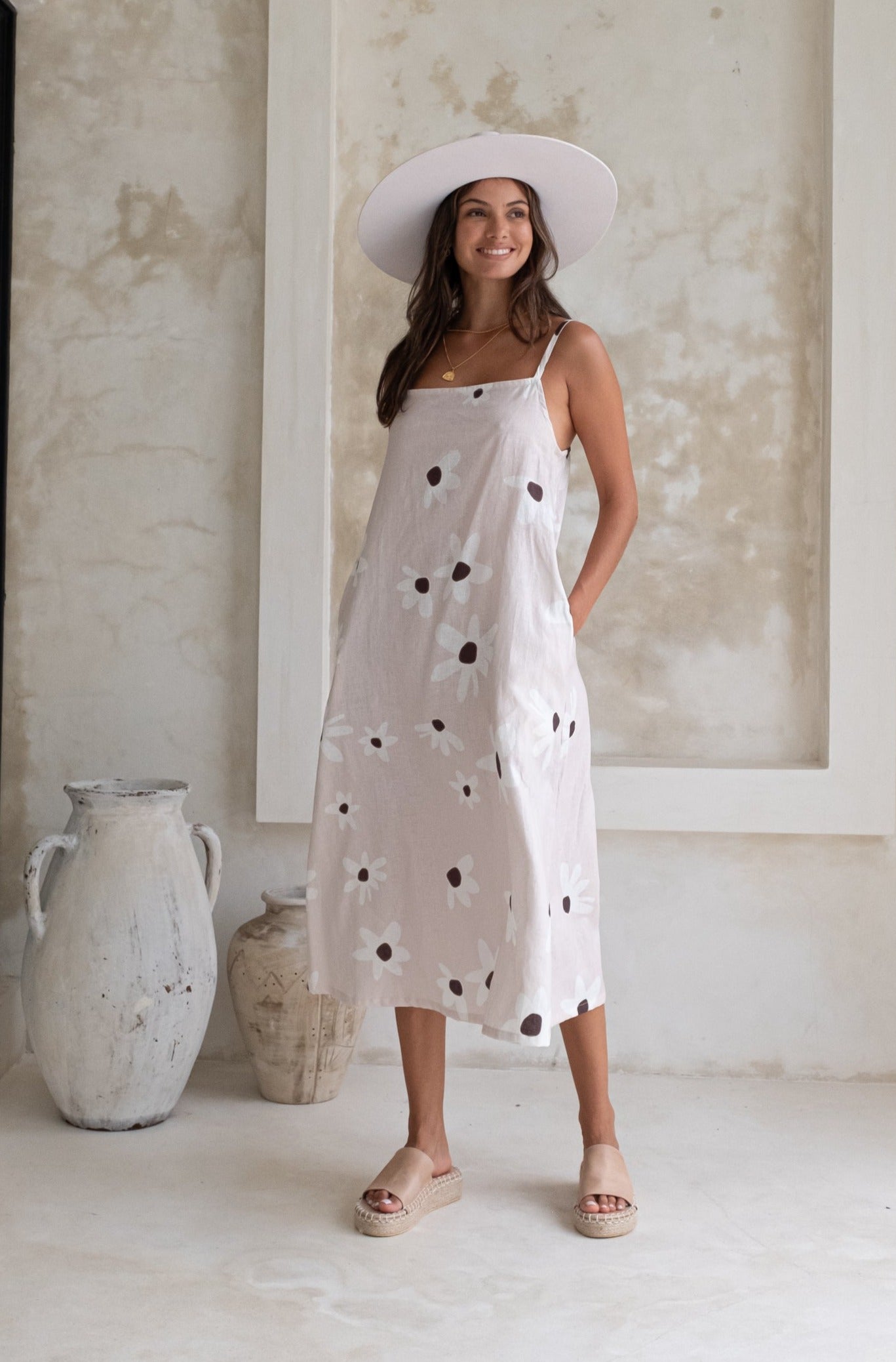 A woman wearing a floral sundress and a wide-brimmed hat, standing in a minimalistic indoor setting with pottery in the background.