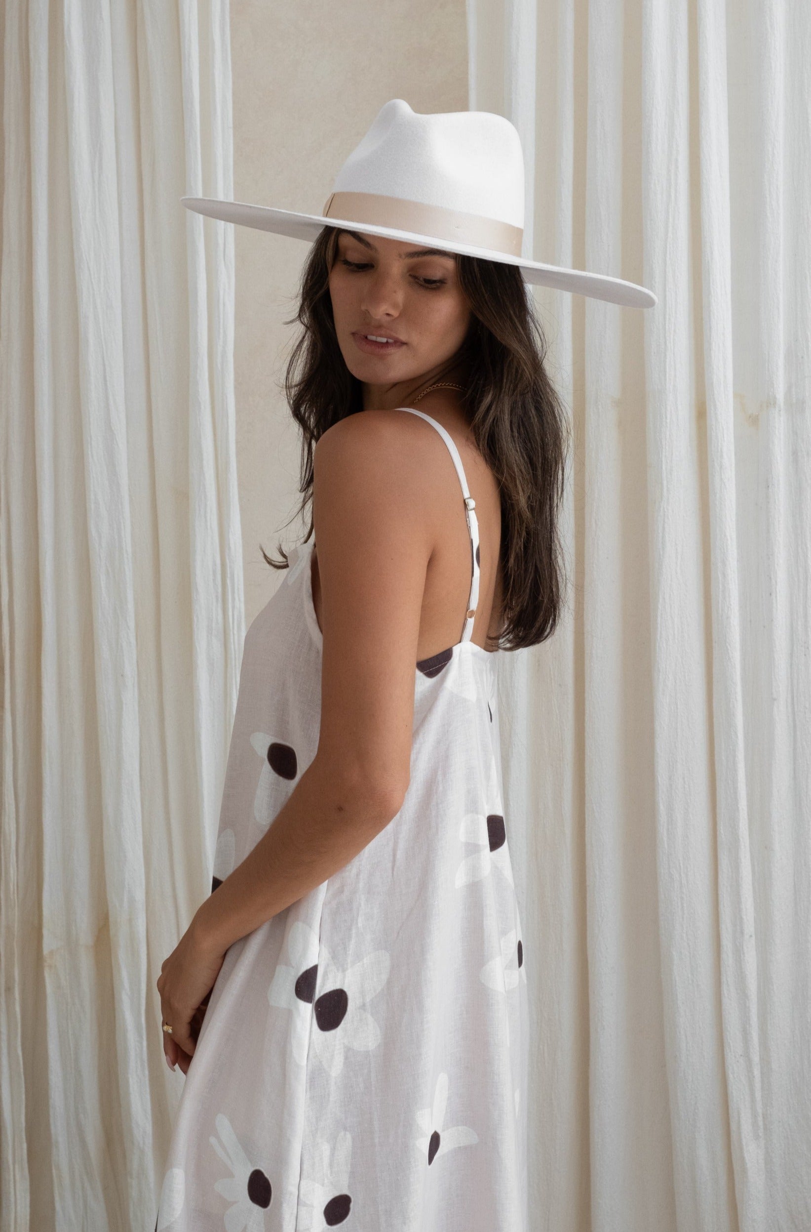 A woman in a floral sundress and white hat, facing sideways, with white curtains in the background.