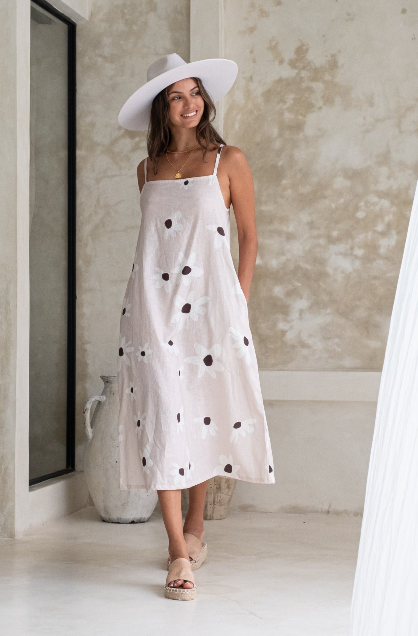 A woman smiling in a floral sundress and white hat, posing in a minimalistic room with pottery and neutral-colored walls in the background.