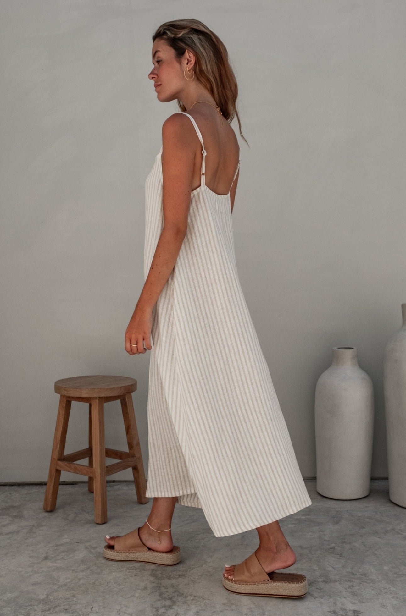 A woman with blonde hair walking in a striped dress, from a side view, placed in a minimalistic indoor setting.