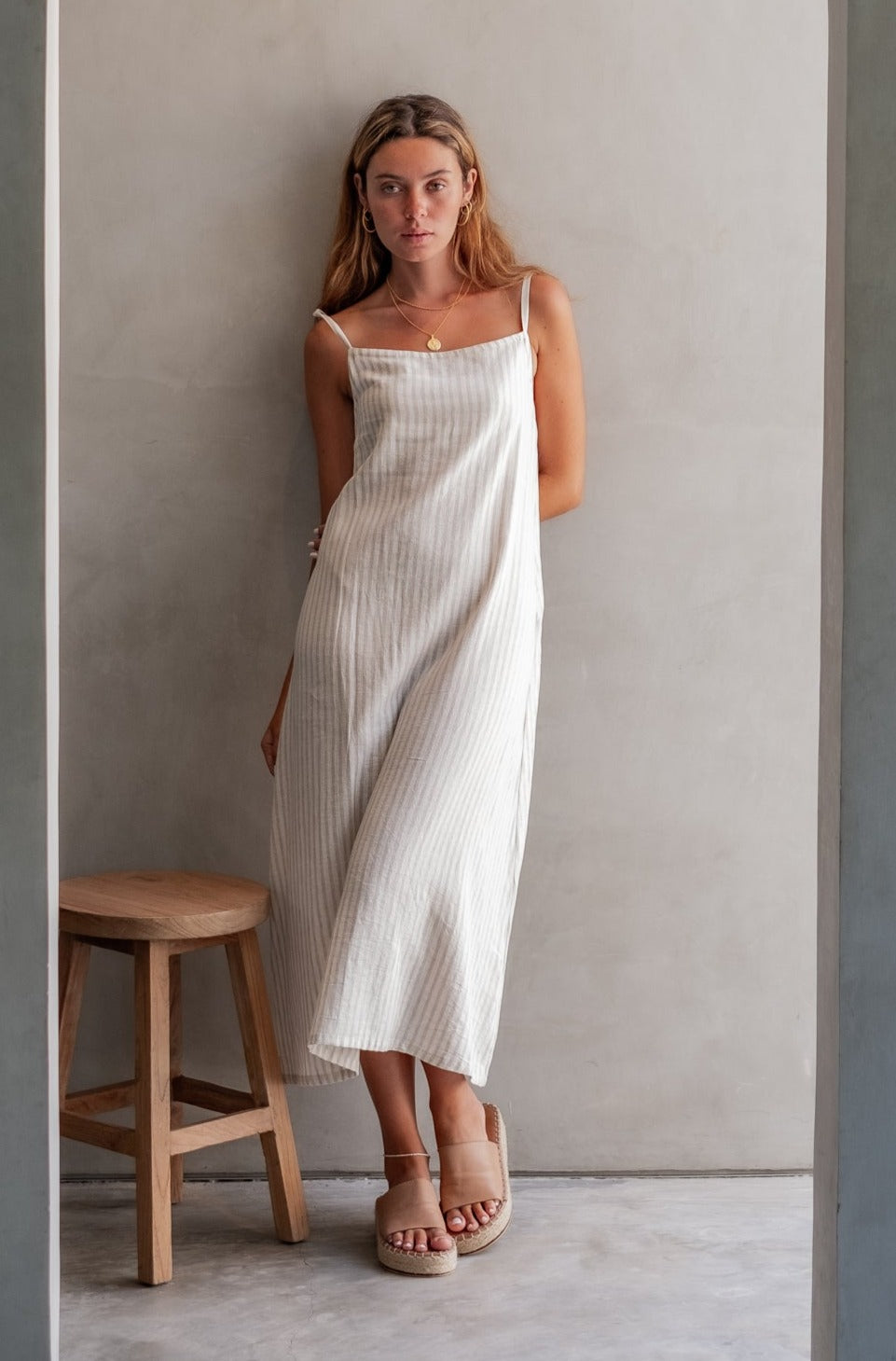 A woman with blonde hair in a striped dress, standing against a wall in a minimalistic indoor setting.