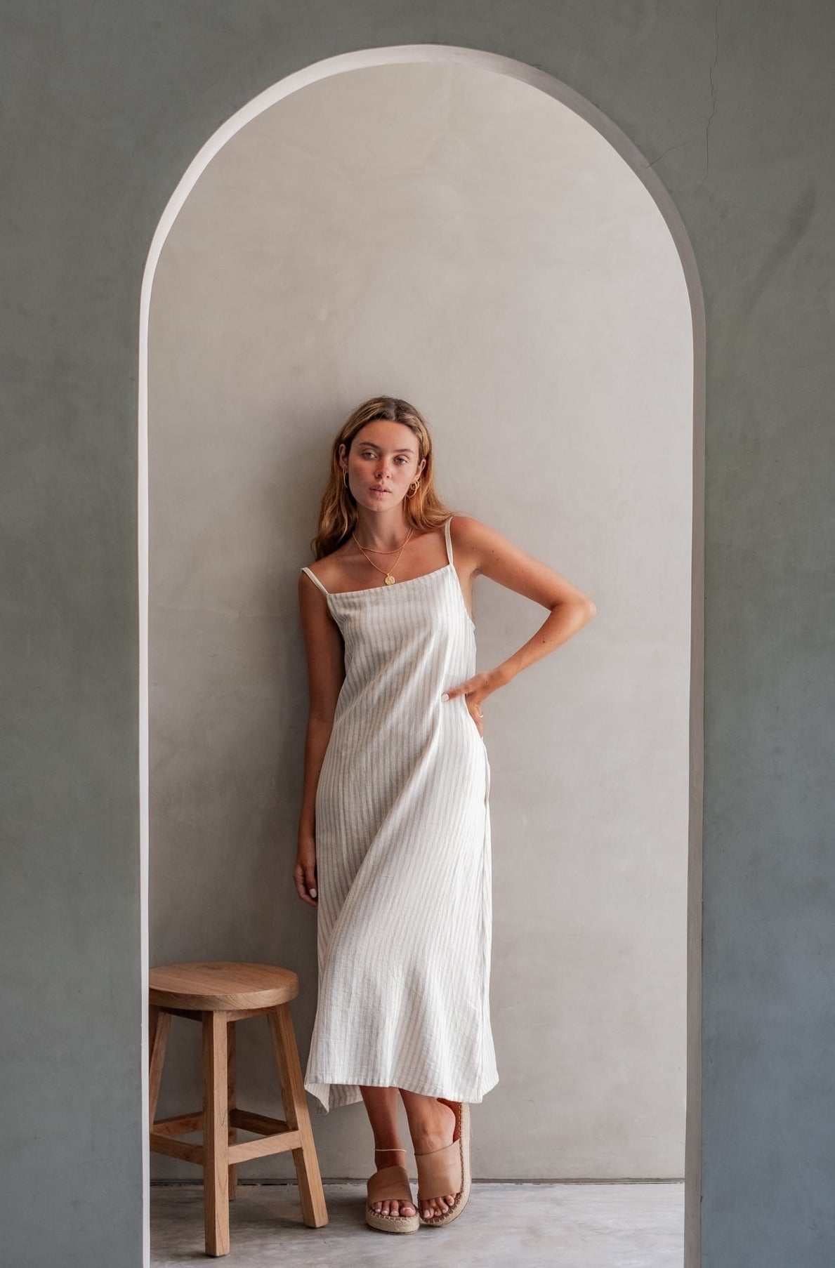 A woman in a striped midi dress, standing in a minimalist space with a wooden stool beside her and an arched doorway