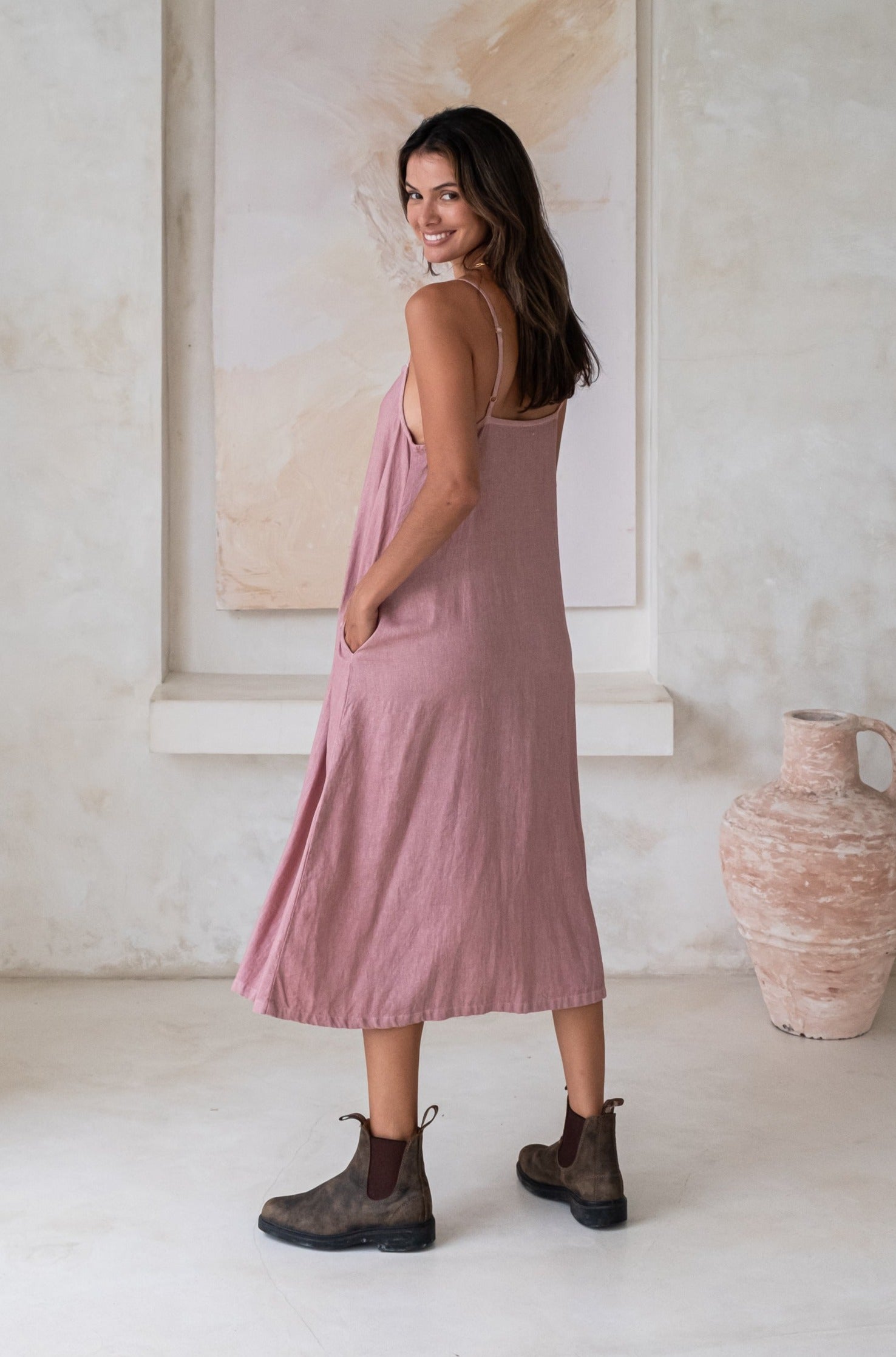 A woman wearing a pink midi dress and green boots while looking over her shoulder in a minimalistic outdoor setting.