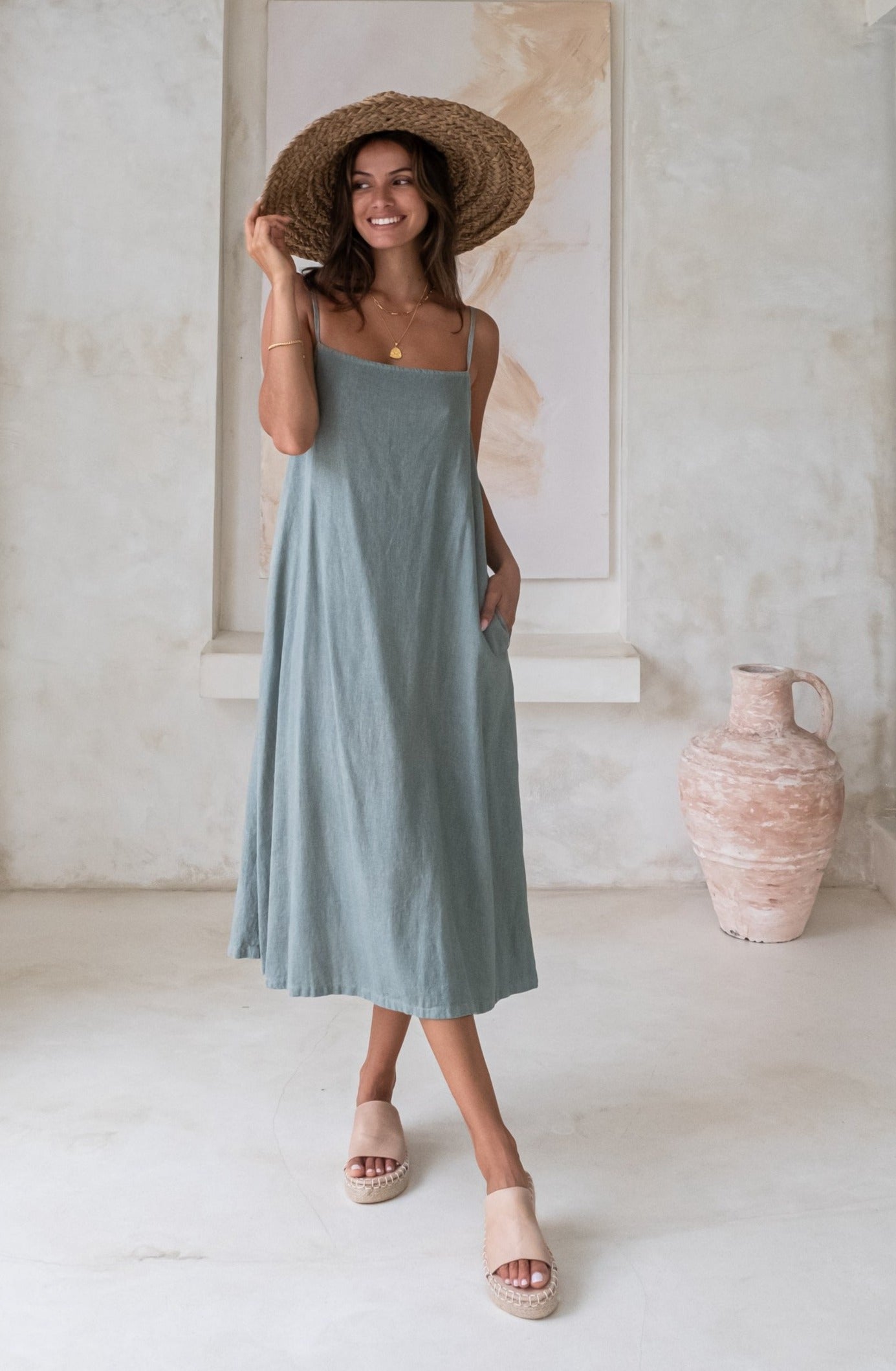 A woman in a light blue sundress and wide-brimmed straw hat, standing in a minimalistic room with neutral-colored decor and pottery in the background.