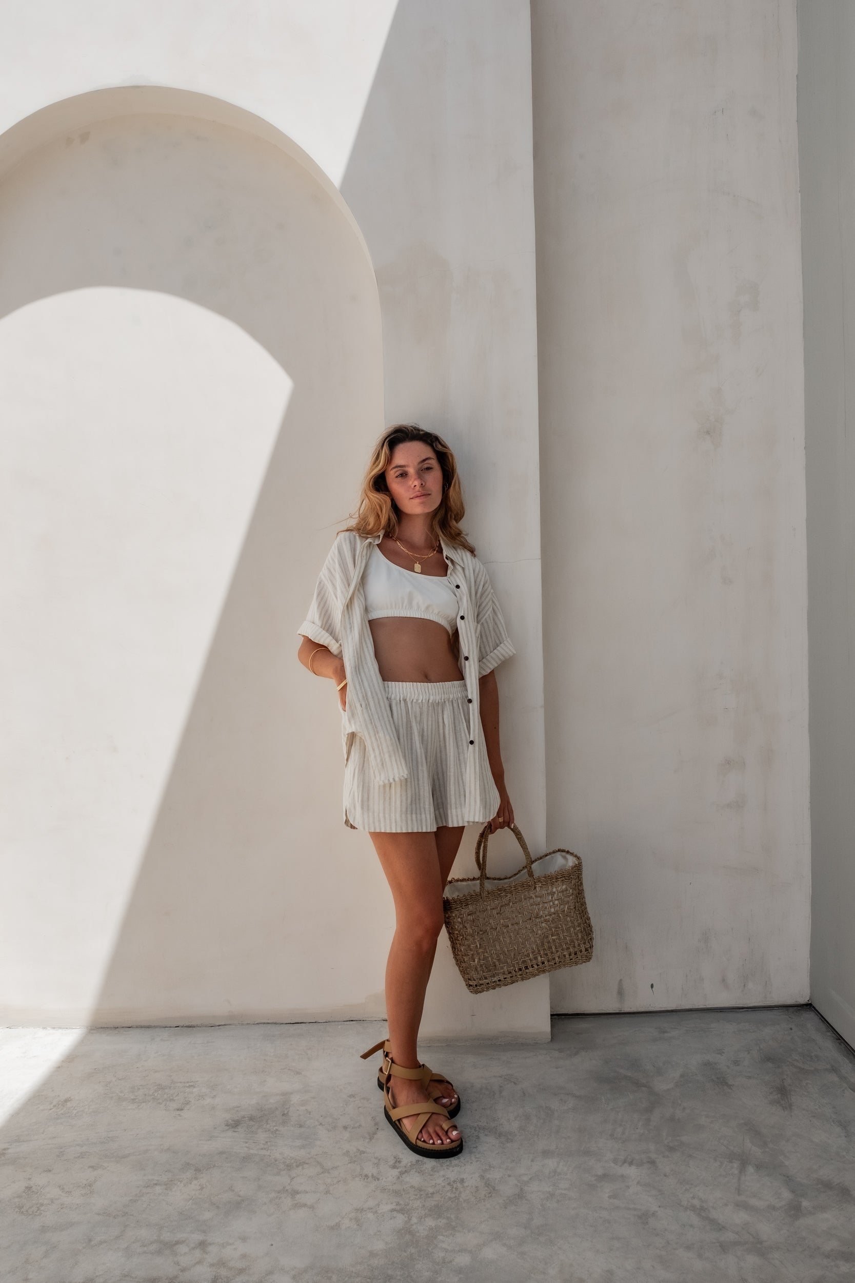 A woman in light pink pajama shorts and a matching loose shirt holding a wicker bag while standing against a minimalistic light pink wall