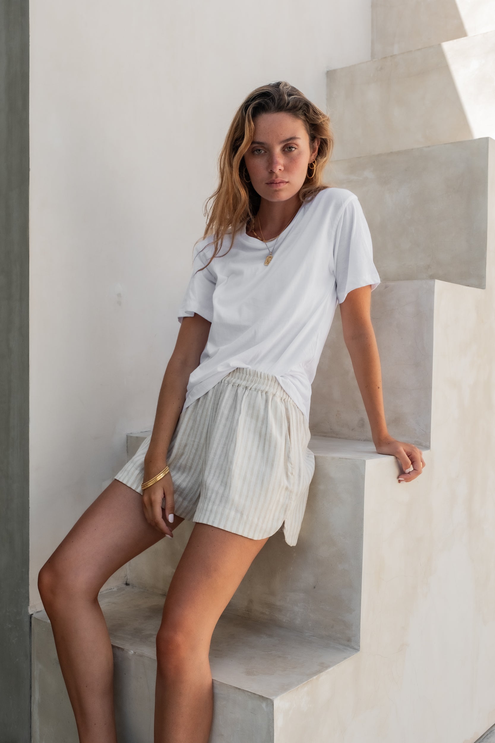 A woman standing against a stair in light pink striped pajama shorts with a white tee and golden jewellery