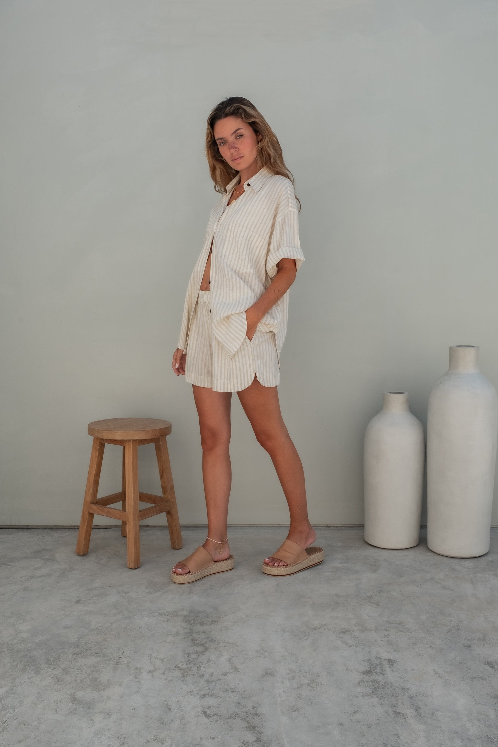 A woman wearing light pink pajama shorts and a matching loose shirt stands against a grey wall with one hand resting in her pockeet looking to the side.