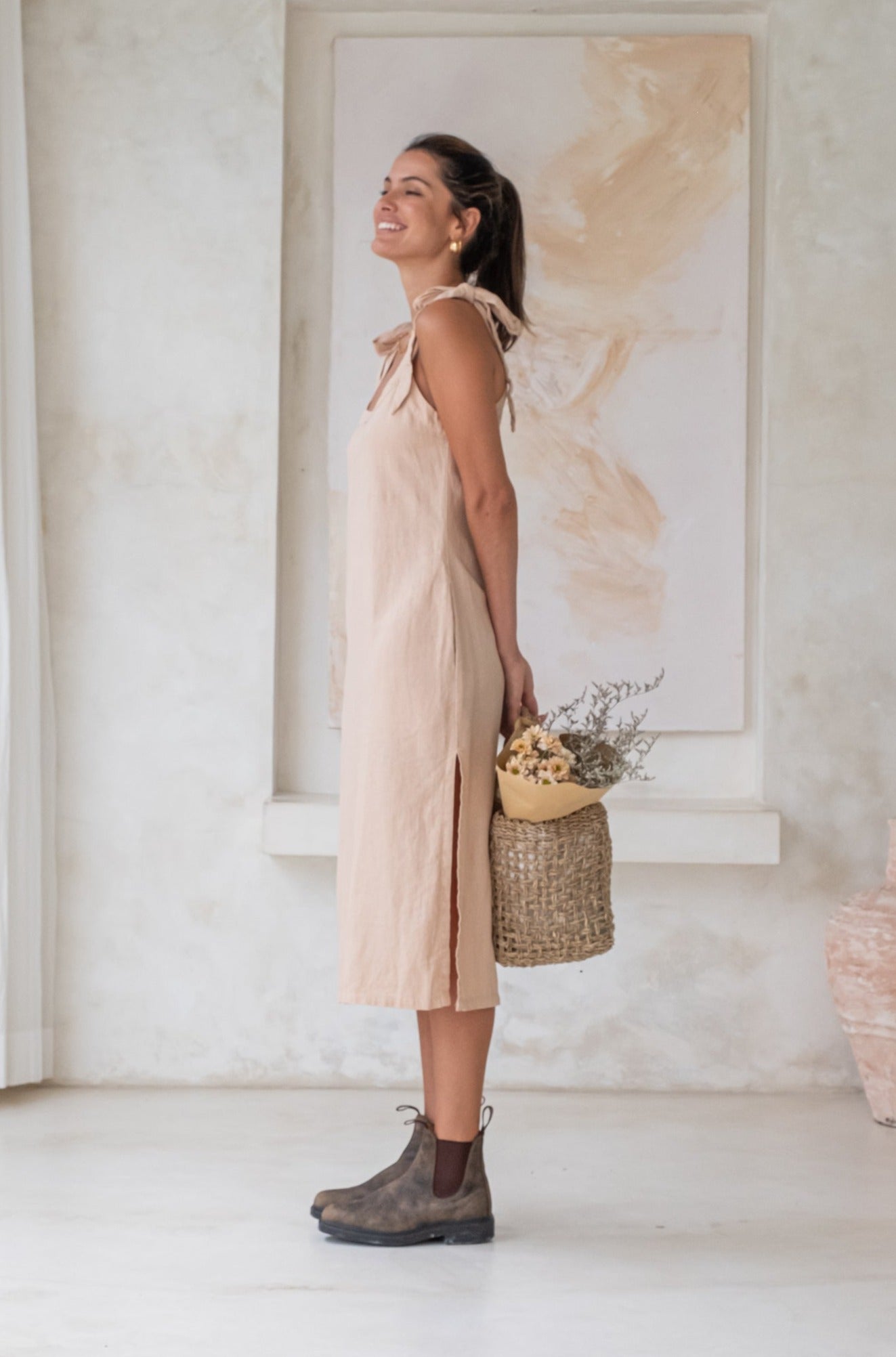 A woman in a light beige midi dress with tied straps and brown Chelsea boots stands in profile, holding a woven basket with flowers, and smiling in a minimalist room.