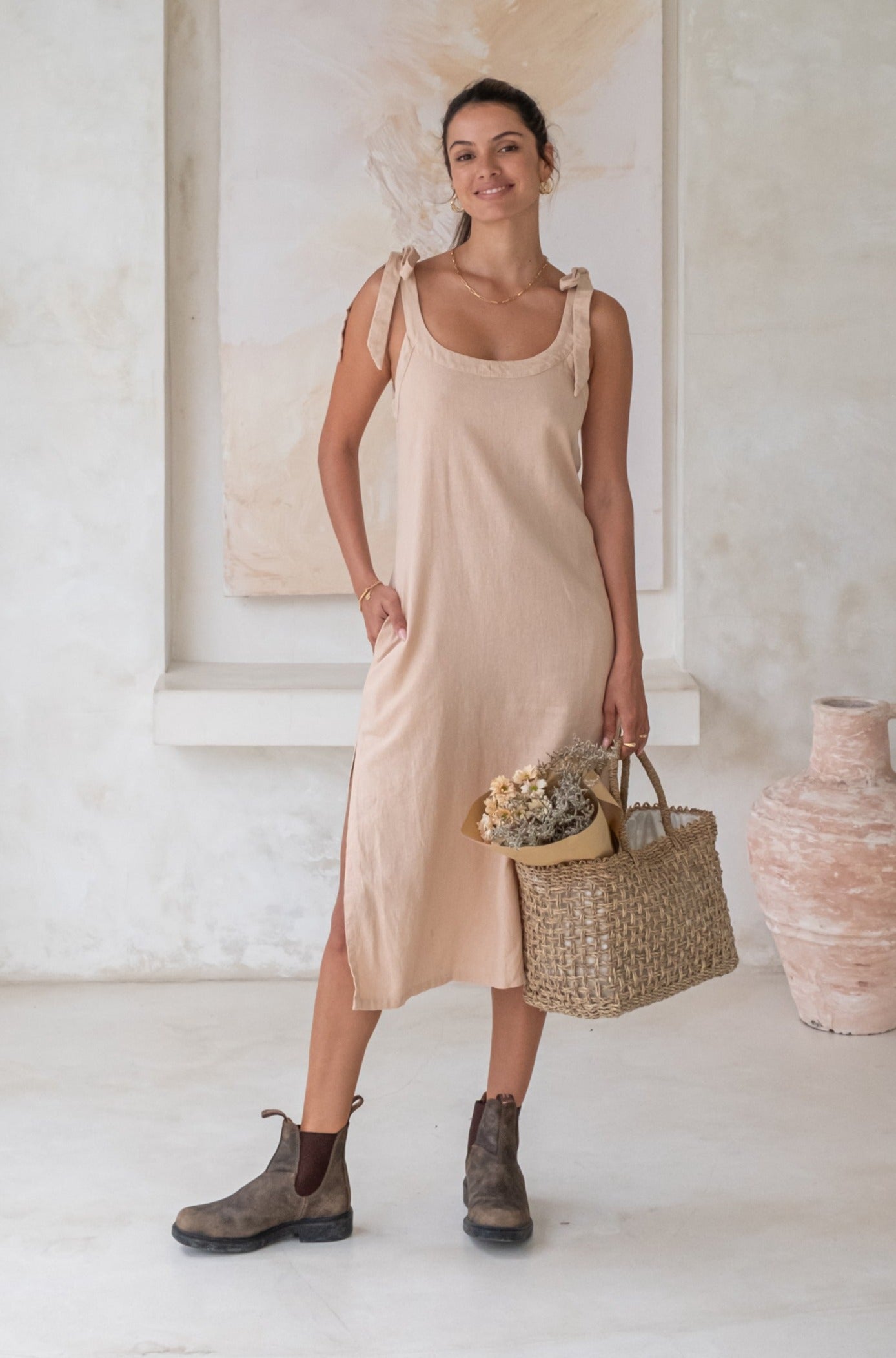A woman in a light beige midi dress with tied straps, brown Chelsea boots, and holding a woven basket with flowers, stands in a minimalist room.