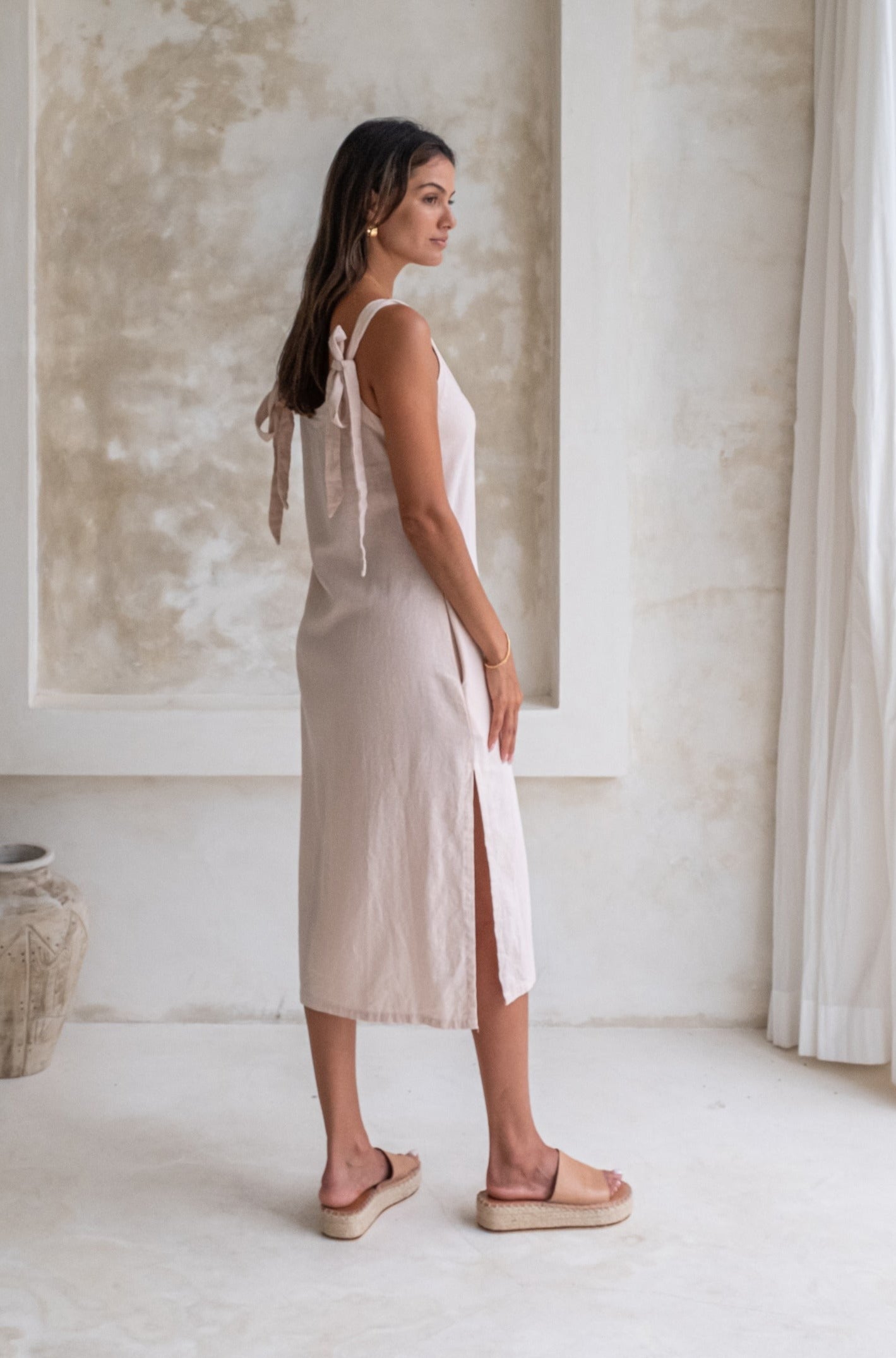 A woman in a light beige midi dress with tied straps and beige espadrille sandals stands in a minimalist room, looking to the side.