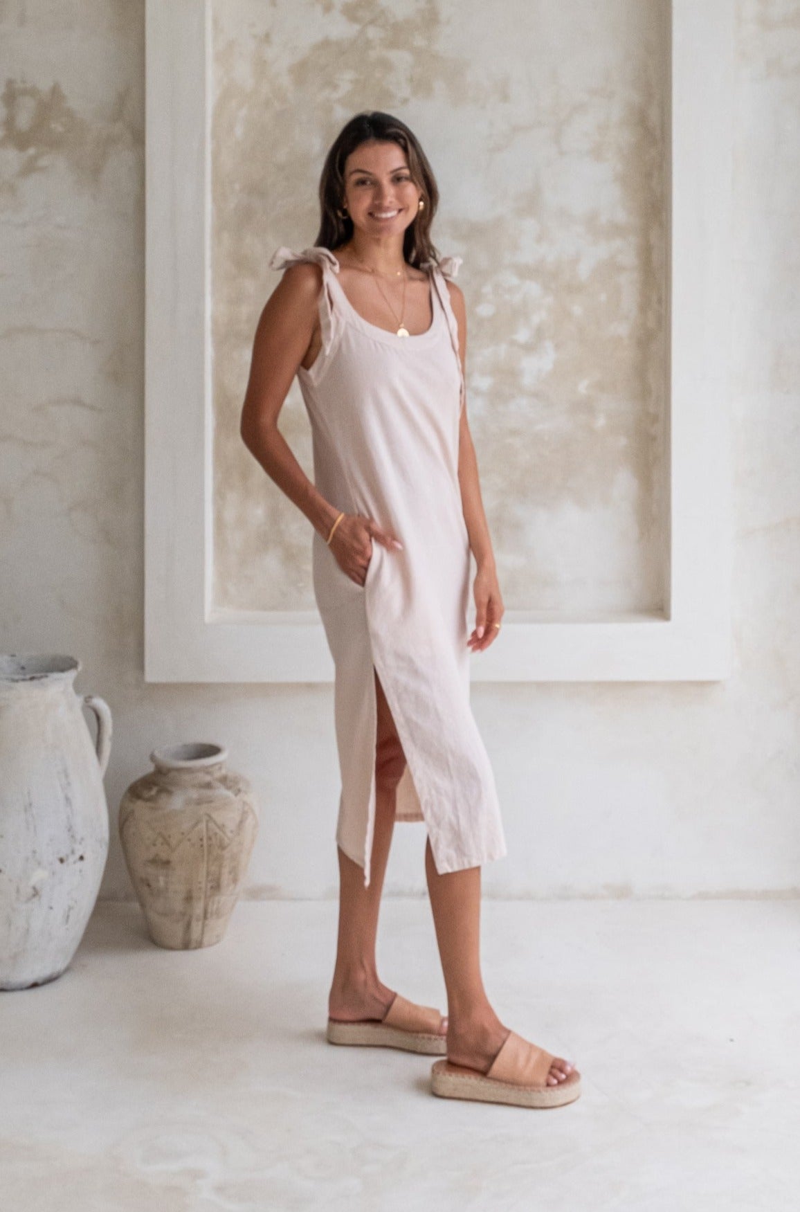 A woman in a light beige midi dress with tied straps and beige espadrille sandals stands in a minimalist room, smiling with her hands in her pockets.