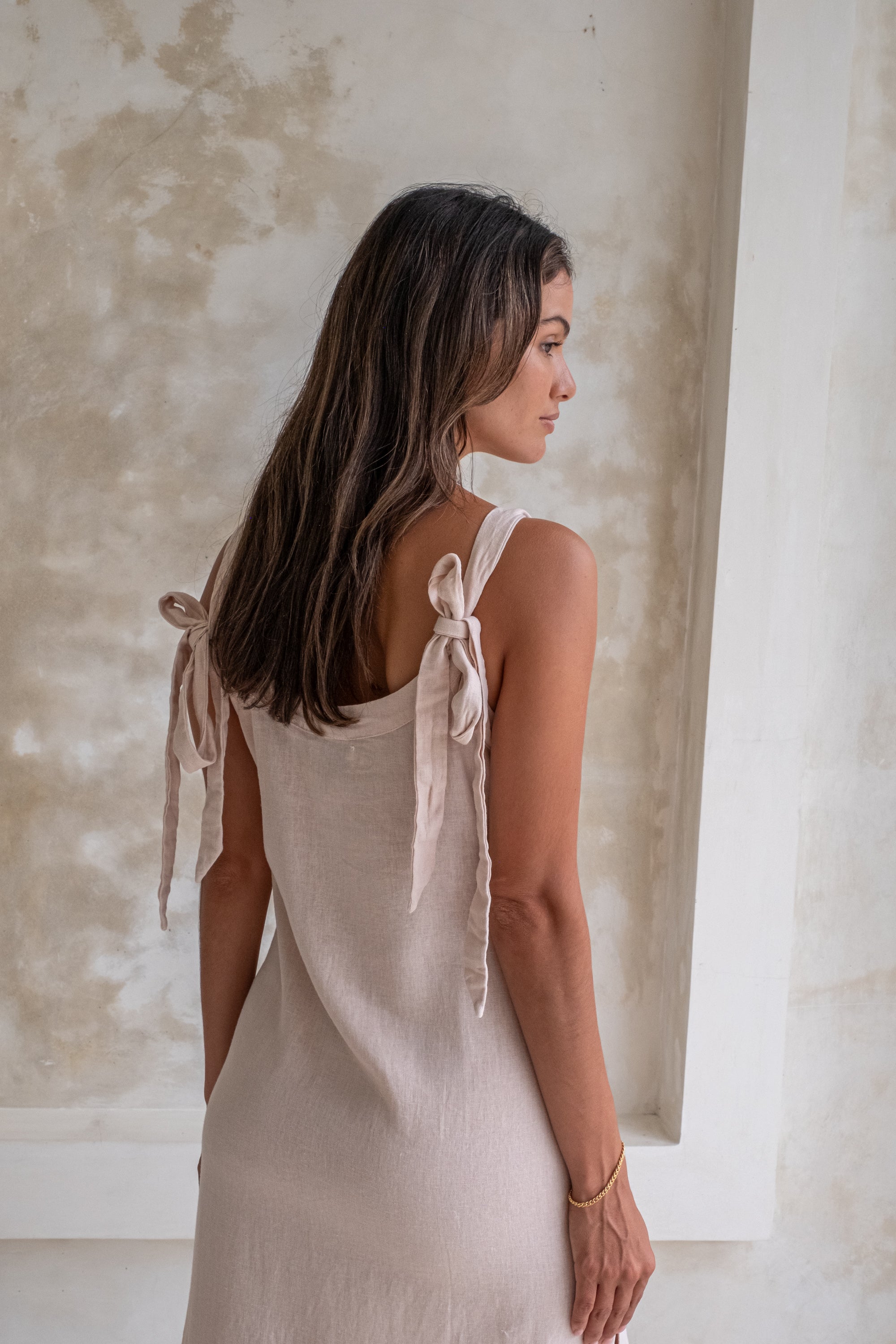 A woman in a light beige midi dress with tied straps, shown from the back, stands in front of a textured wall.