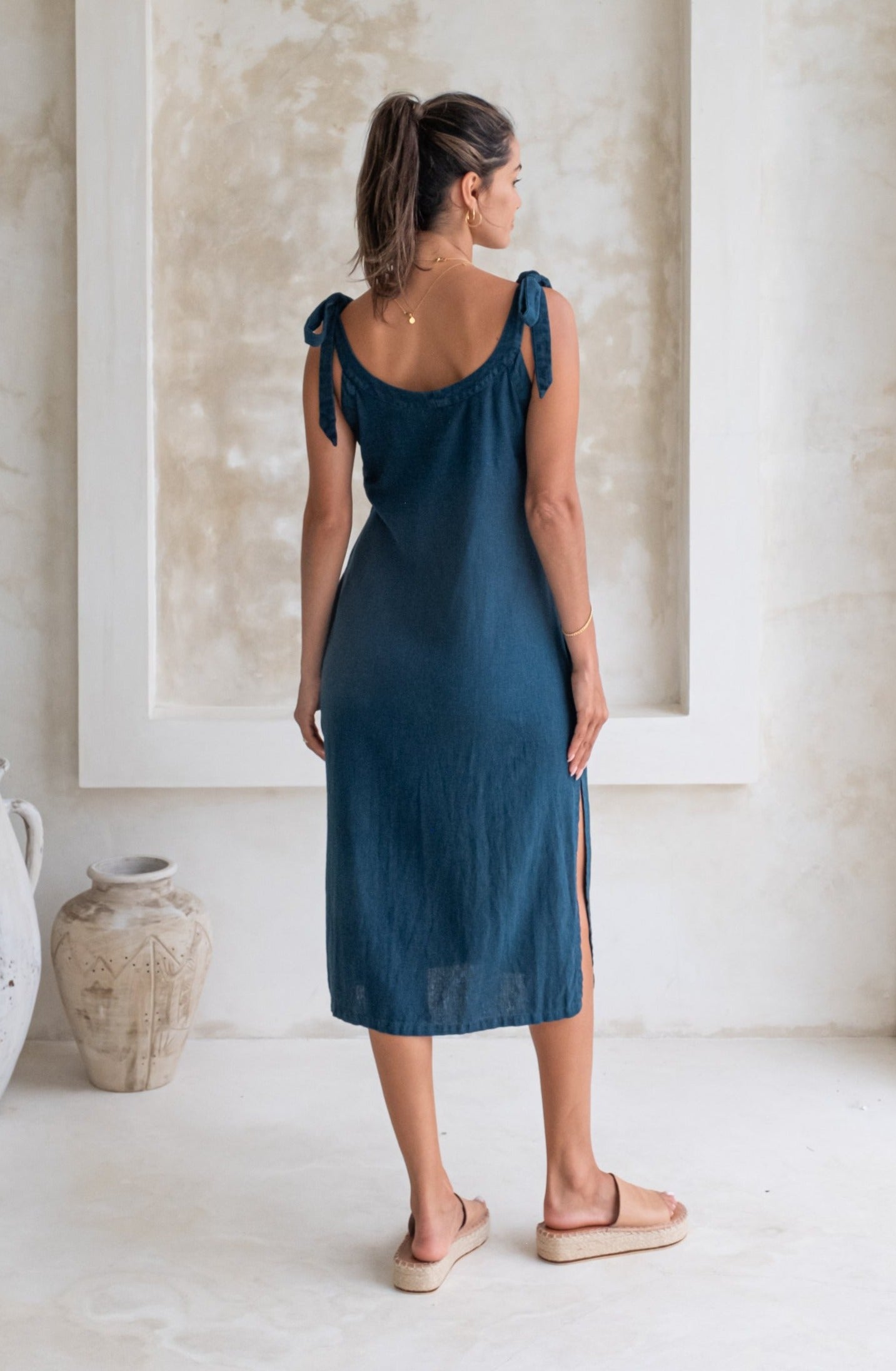A woman in a dark blue midi dress with tied straps and beige espadrille sandals stands in a minimalist room, shown from the back.
