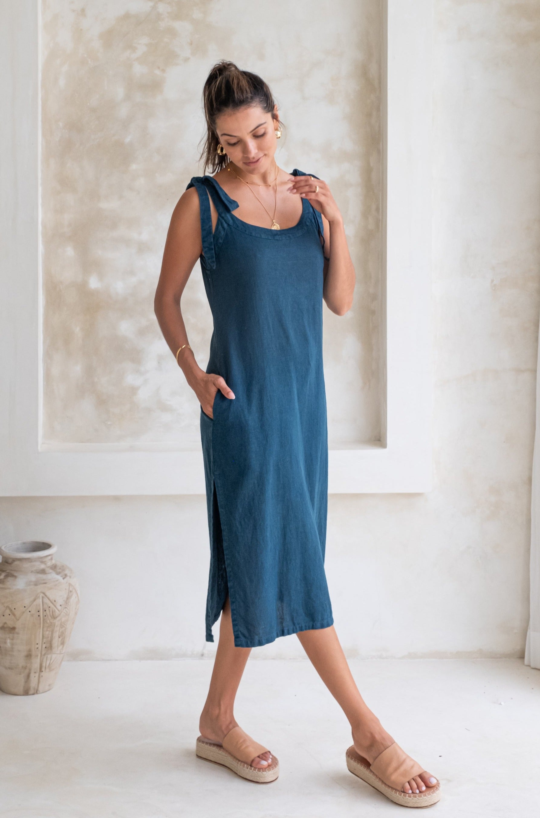 A woman in a dark blue midi dress with tied straps and beige espadrille sandals stands in a minimalist room, looking down and smiling.